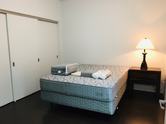 View of a bedroom, mattress set, a small brown side table, a lamp, closet.