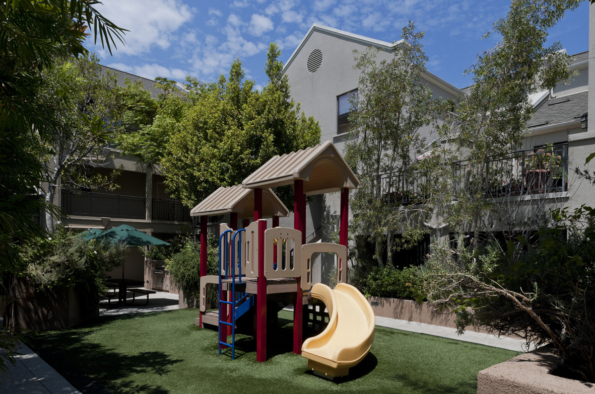 View of an small outdoor pllayground. From this angle, a slide and climbing ladder is visible. Thr ground is short grass. Plants and trees surround the area. Three story gray building is in the background.