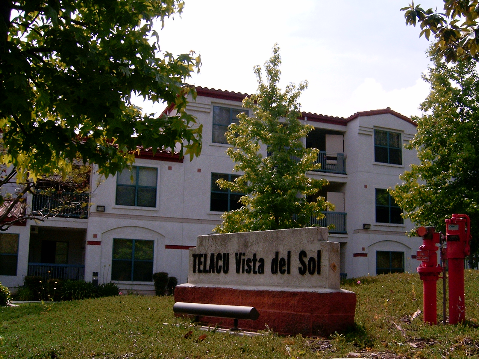 View of a concrete and bricks stone with the building's name "Telacu Vista del Sol", side of the building with multiple windows some with balconies, trees around it, Fire hydrants markers.