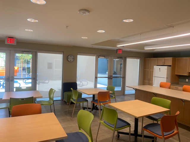 View of a community room, small wood tables with colorful chairs, a white refrigerator, a clock in the wall.