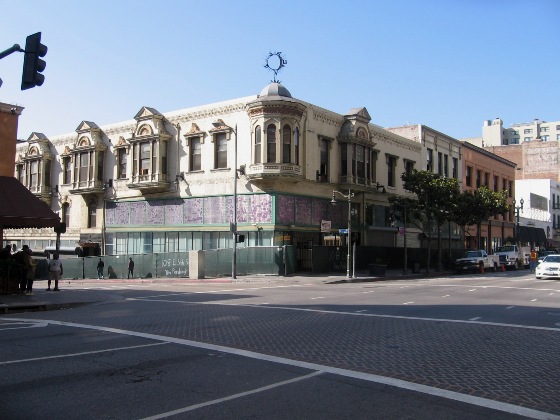 Three story vintage style building on the building of a corner. Green construction fencing surrounds the building. A circular small monument sits on top of the building.