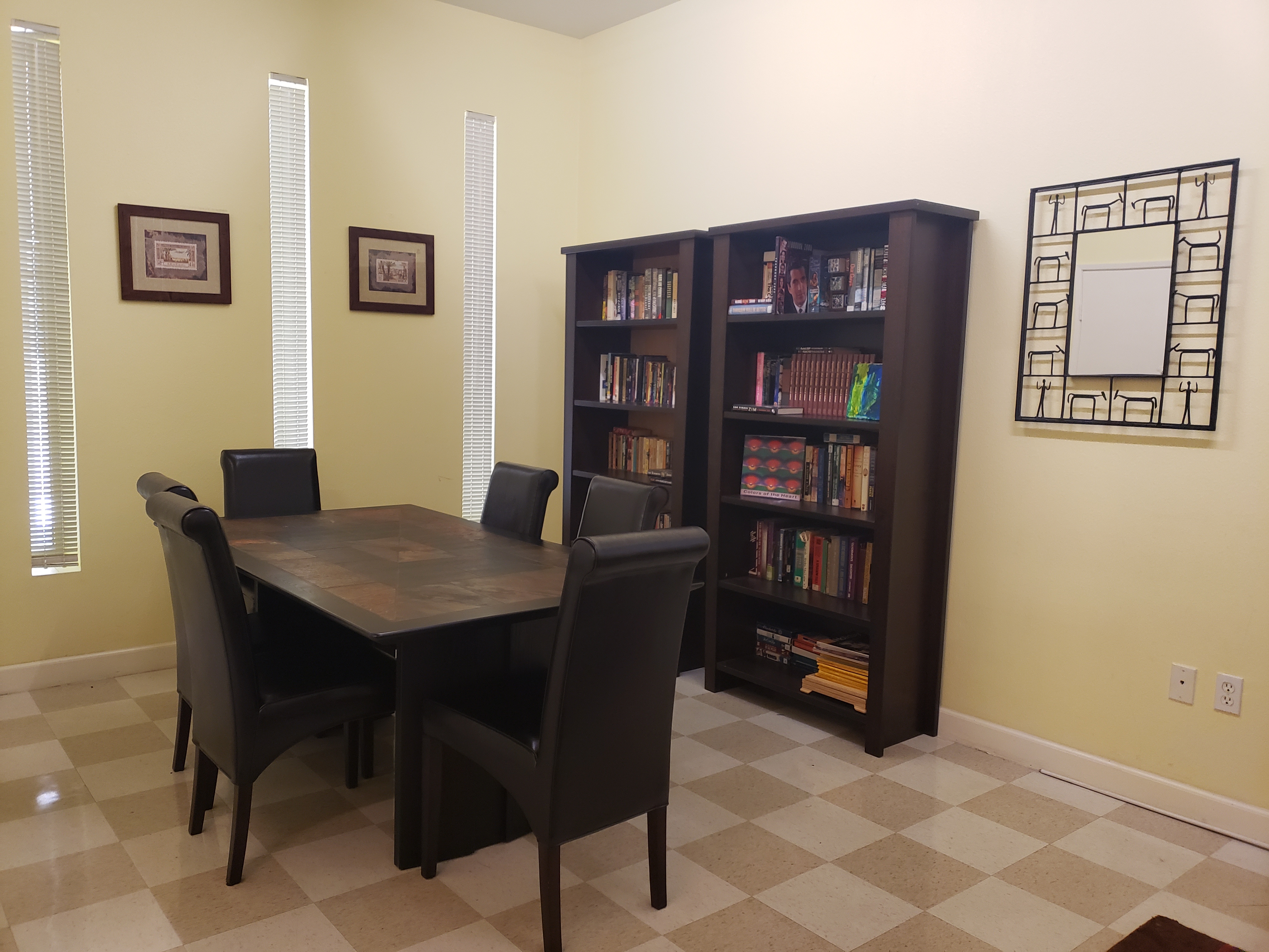 Right side of a room, a brown square table with six brown leather chairs, two brown tall shelves full of books, three decorative frames on the wall, windows with horizontal blinds.