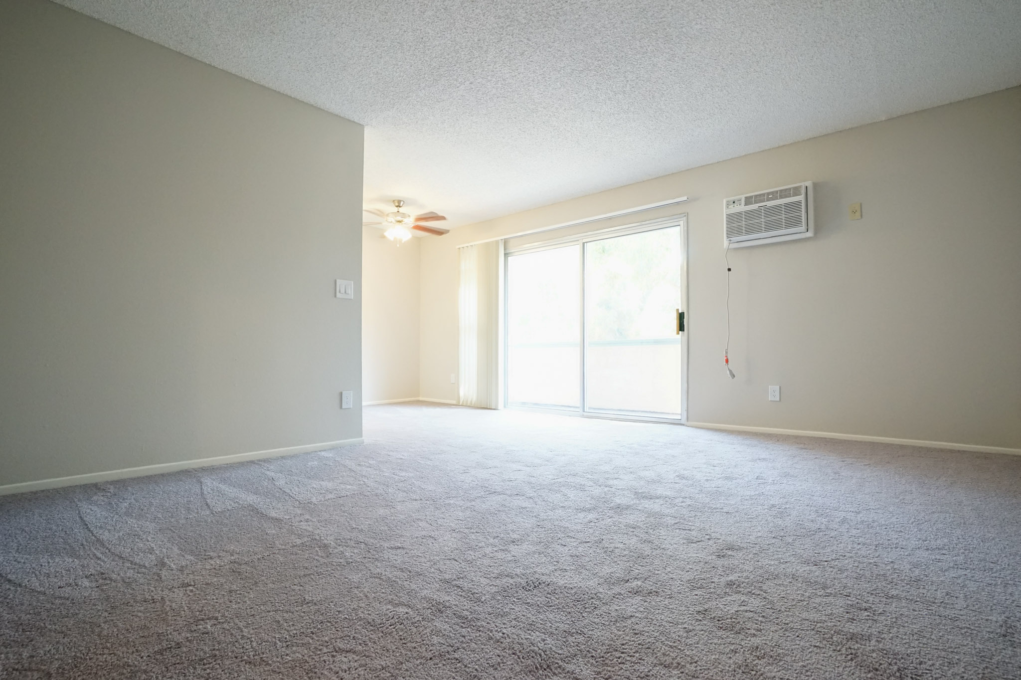 Living room with a large sliding door. There is a ceiling fan and an air conditioner mounted on the wall. Floor is carpeted.