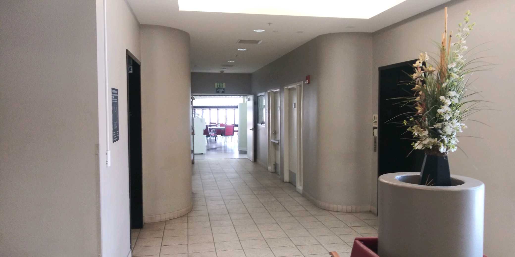 Hallway view of of lobby area. There are two elevators across from each other. There is a large lounging room pictured with multiple table sets in the distance.
