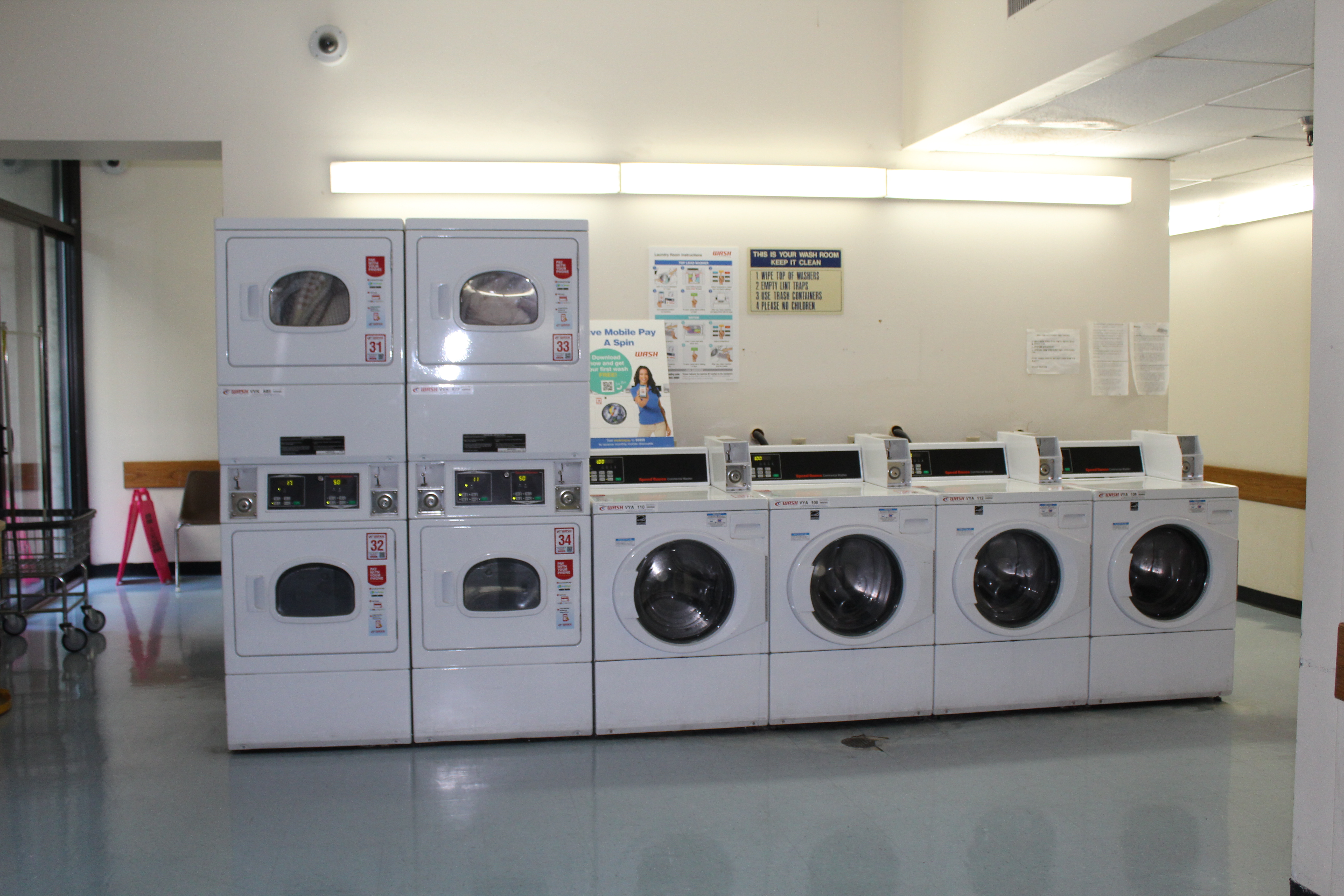 Interior view of a laundry room for Angelus Plaza 1. 4 white washers with front circular windowed doors. Two rows of white dryers, 4 total