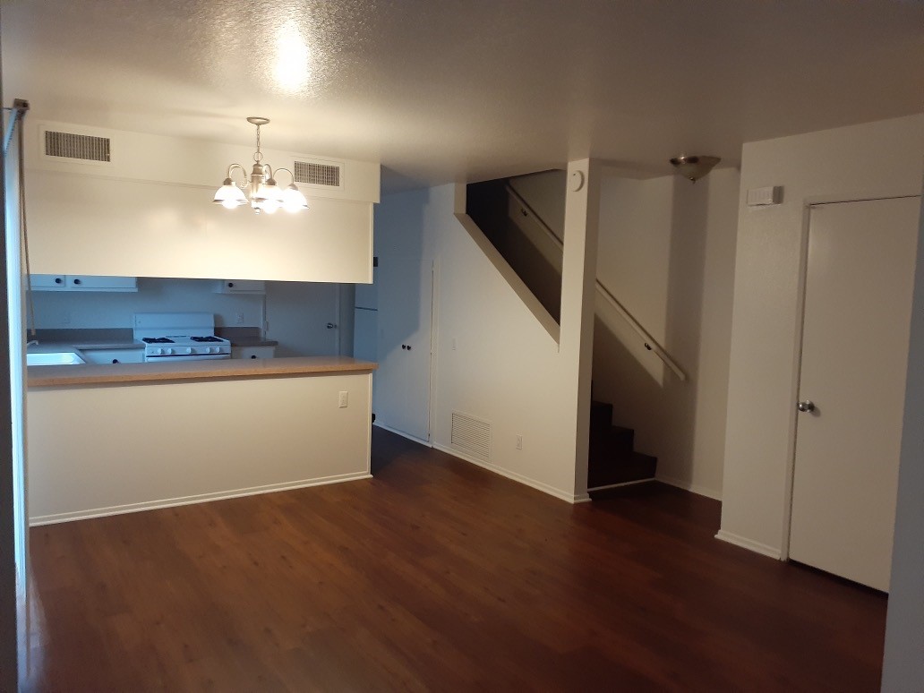 View of a empty unit, part of living room and part of the white kitchen, brown laminate floors, stairway on the right side, a chandelier.