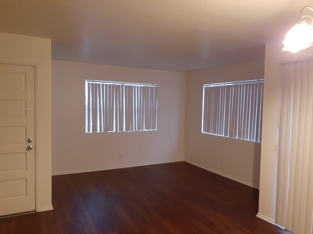 View of an empty living room, brown laminate floors, two windows with white vertical blinds, white walls.