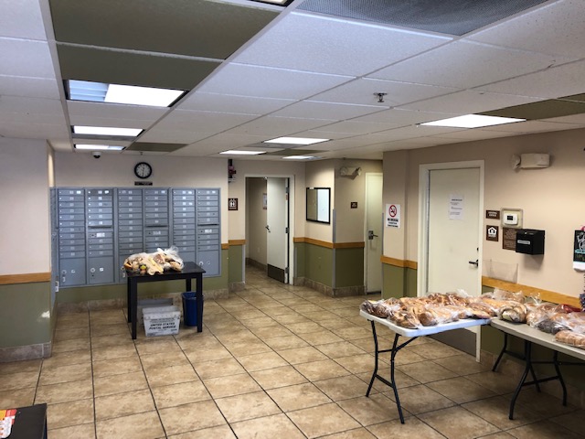 Front view of common area, two portable tables with many bags of sandwhich bread, on the left side far back against the wall a whole set of gray mailboxes, a clock on the wall on top of the mailboxes, multiple doors, multiple lights.