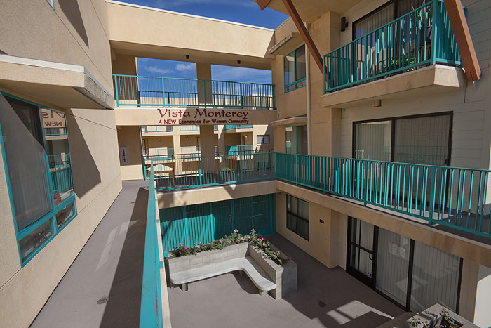 View of the inside of a three story beige building. Unit access is through outdoor passageways with teal railings. Ground floor has a cement bench with plants behind it. Units have large windows.