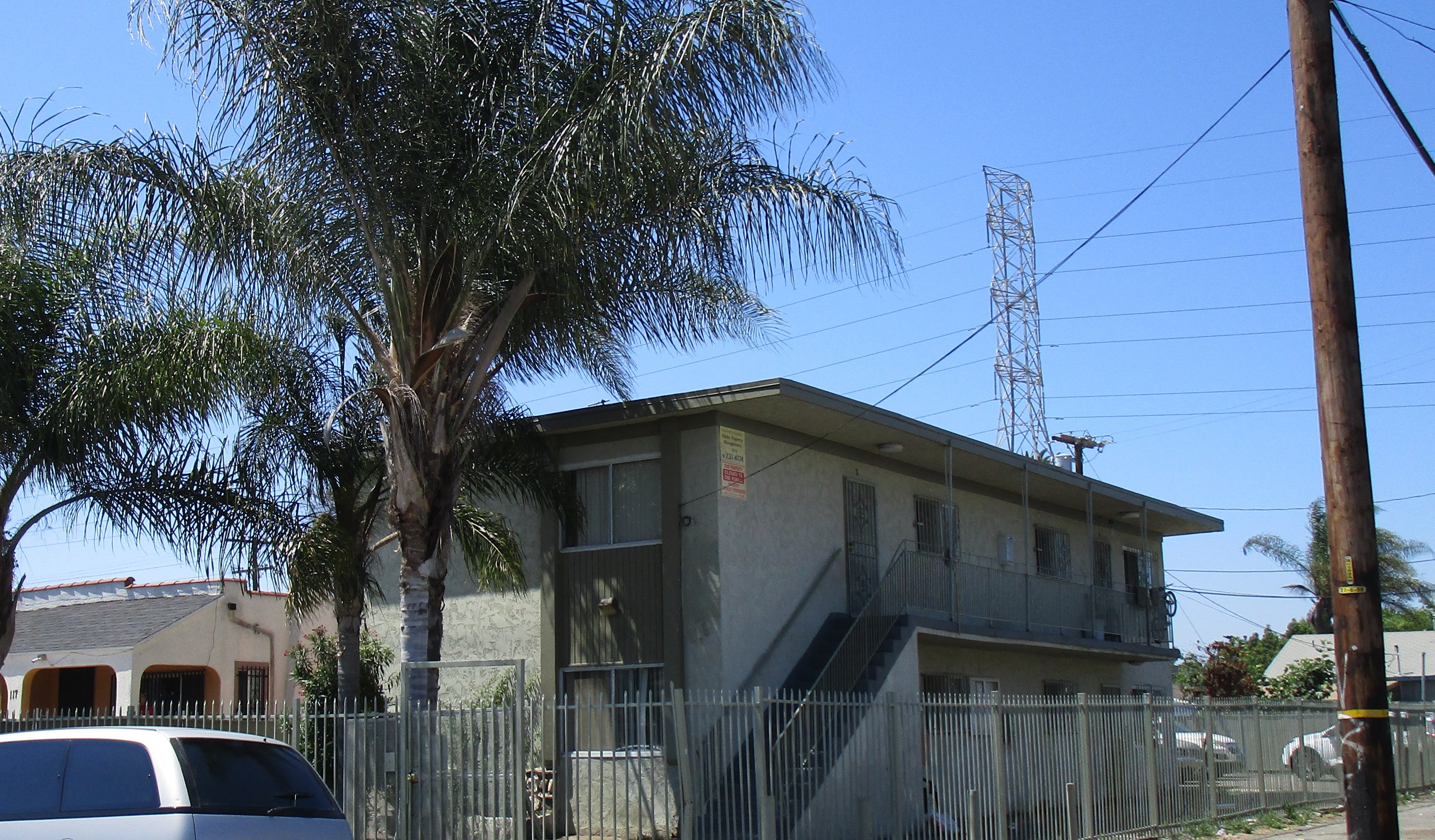 Corner side view of a two story building, multiple gated windows and iron screen doors, all gated, stairs up to the second floor with iron handrails, tall palm tree, light pole and parked cars, power tower in the back by the parking lot.