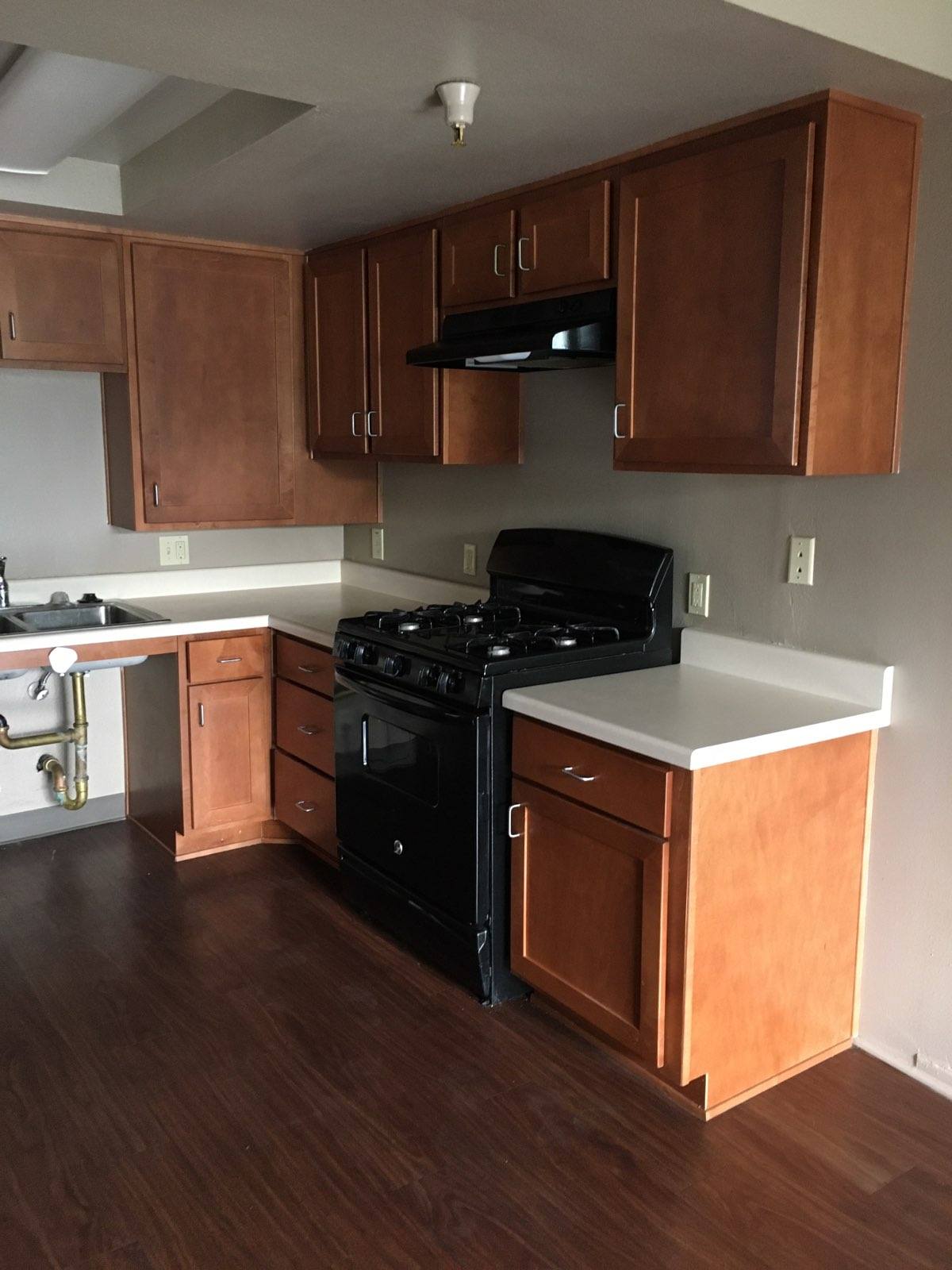 View of upper and lower kitchen cabinets, white countertops, black stove and hood, and wheelchair accessible sink.