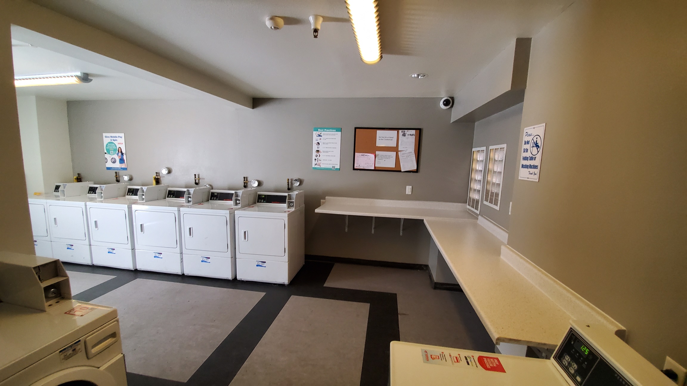 Laundry room with multiple washer and dryer machines. Room has a shelf for folding that is L shapes along the walls. There is a security camera on an upper corner of the room, and posted signs across the walls. Floor area is spacious.