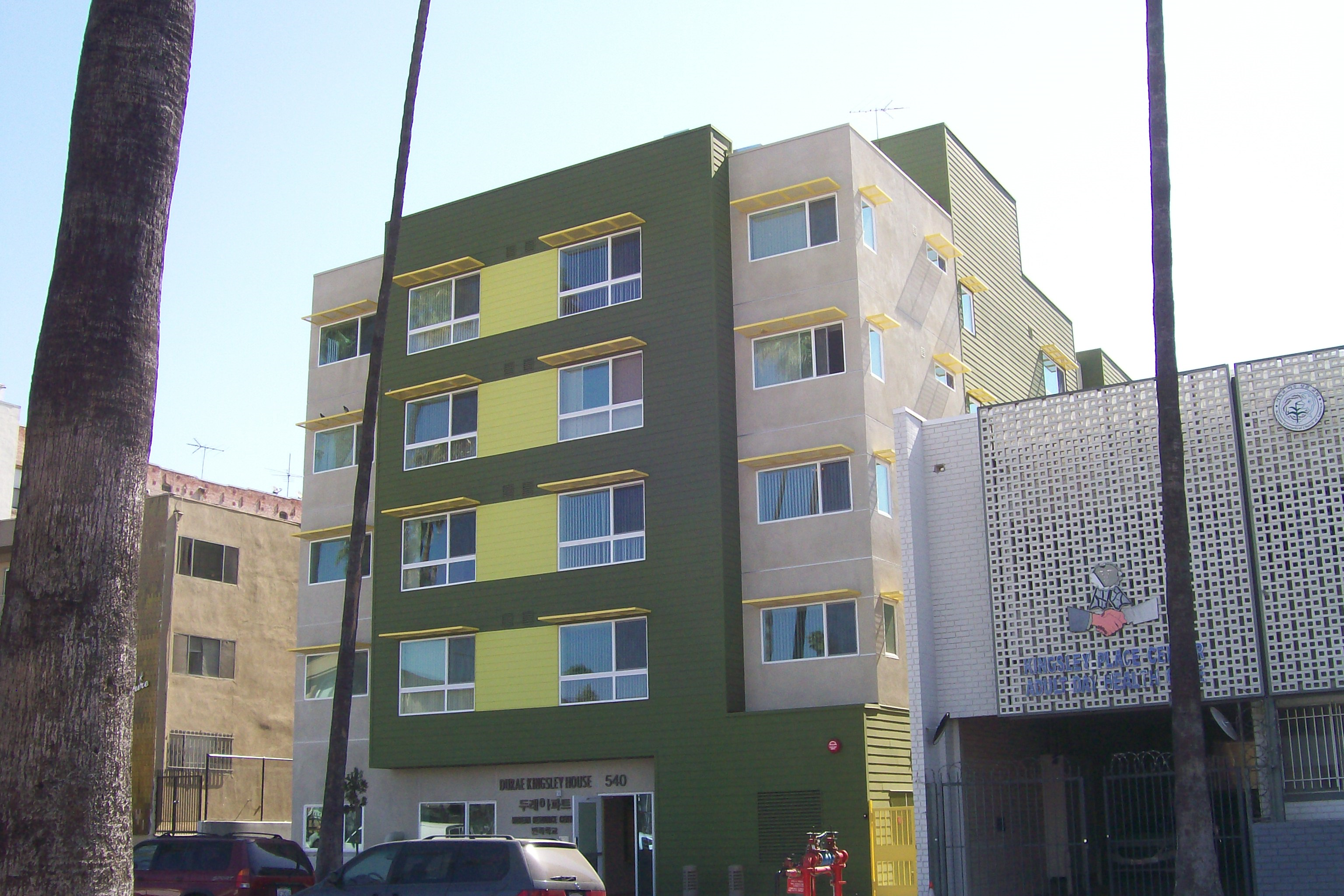 Right side of a colorful four story building, dark green-Lime green and beige colors, multiple windows, parked cars, fire hydrant, palm tree.