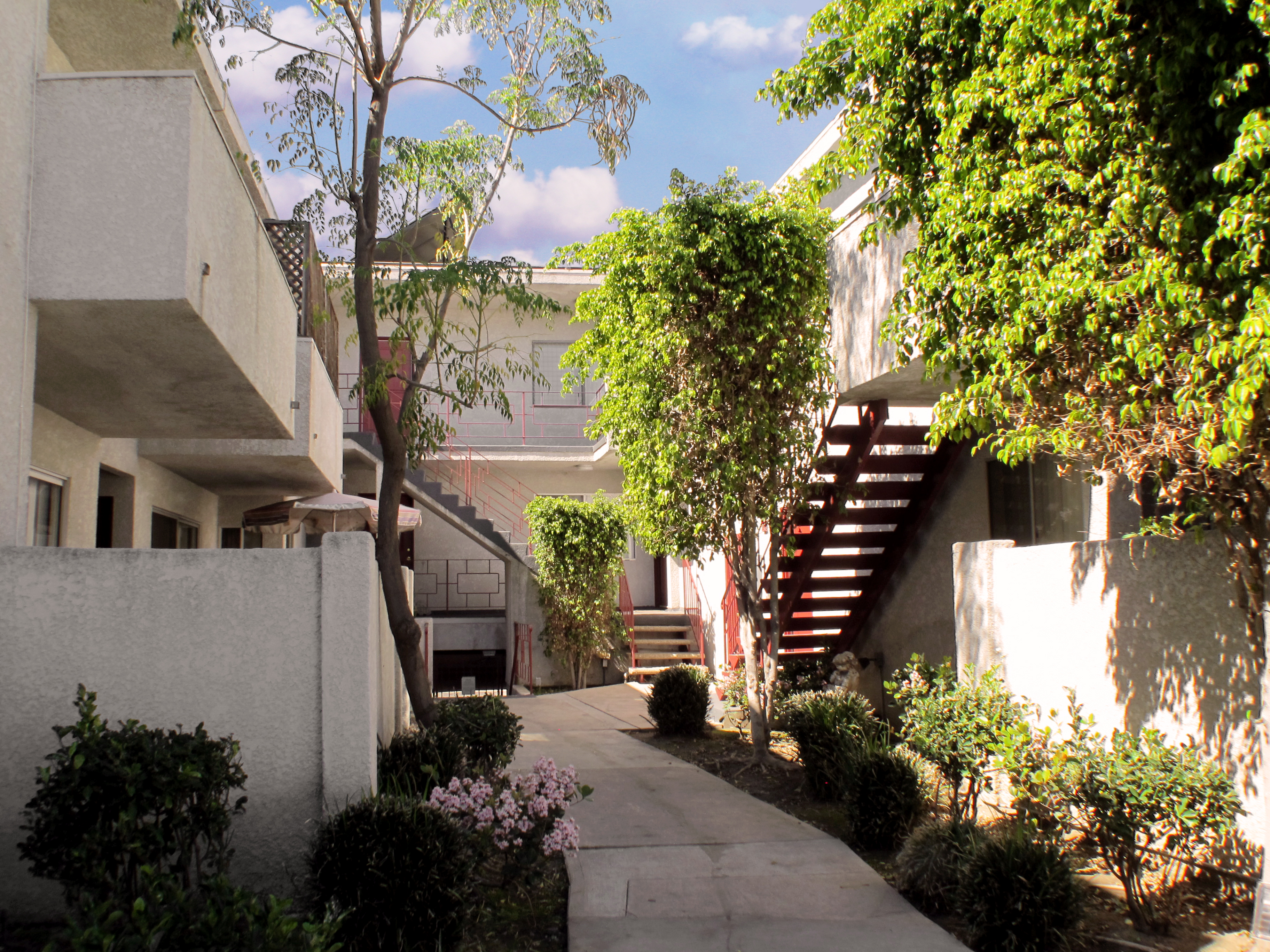 Courtyard of a two story white building. There's a passageway with plants and trees on the sides. There are staircases with handle bars and balconies visible.