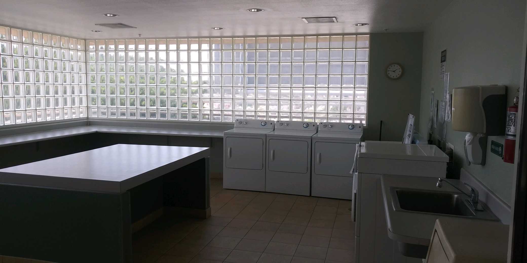 Laundry room with multiple washers and dryers. Center has a folding table. There is a large tiled window on two sides of the wall amd a shelf alongside it. On the adjacent wall there are laundry room rules and signs posted. Thre is a sink, soap dispenser,