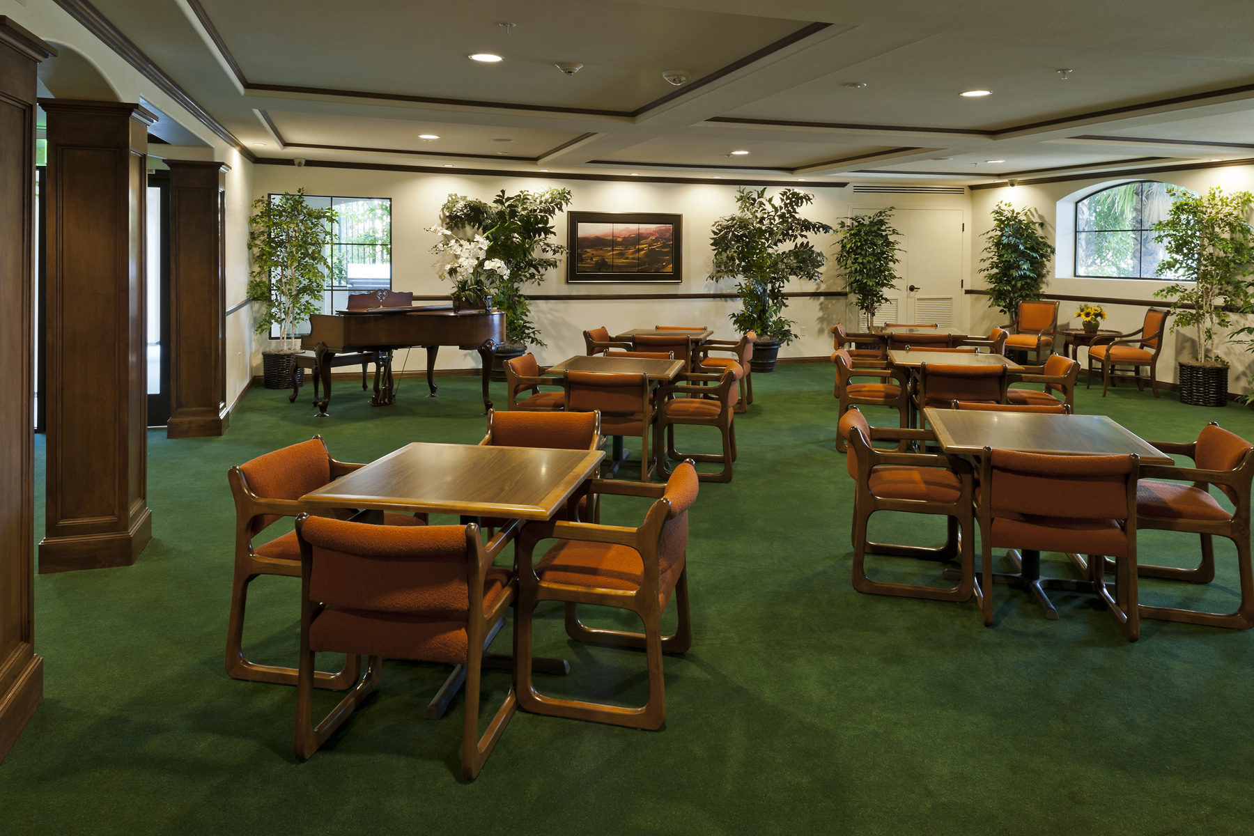 Canby woods housing community room. large wood with green carpet, several brown wood seating and tables, grand piano, plants throughout room, and painting on wall.