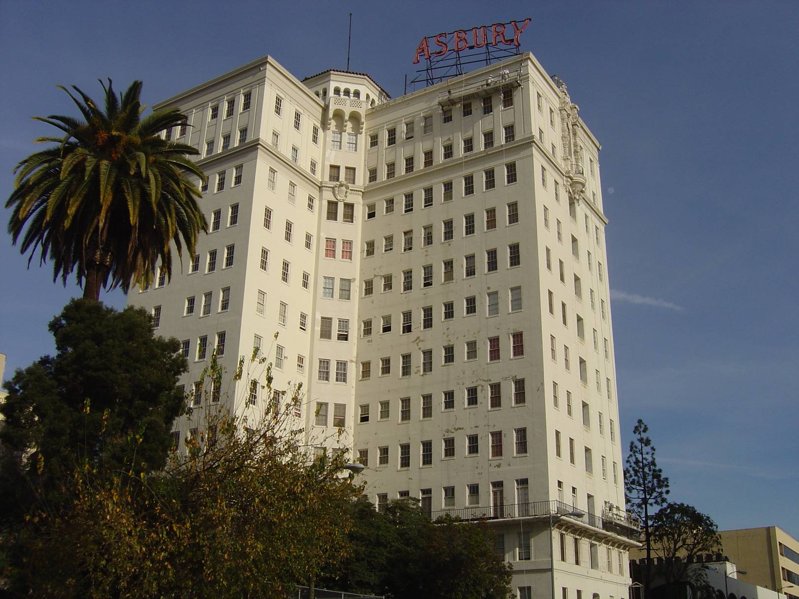 Large twelve story old fashioned building. Building color is of a light brown shade. Rooftop contains a large red sign that reads "Asbury".
