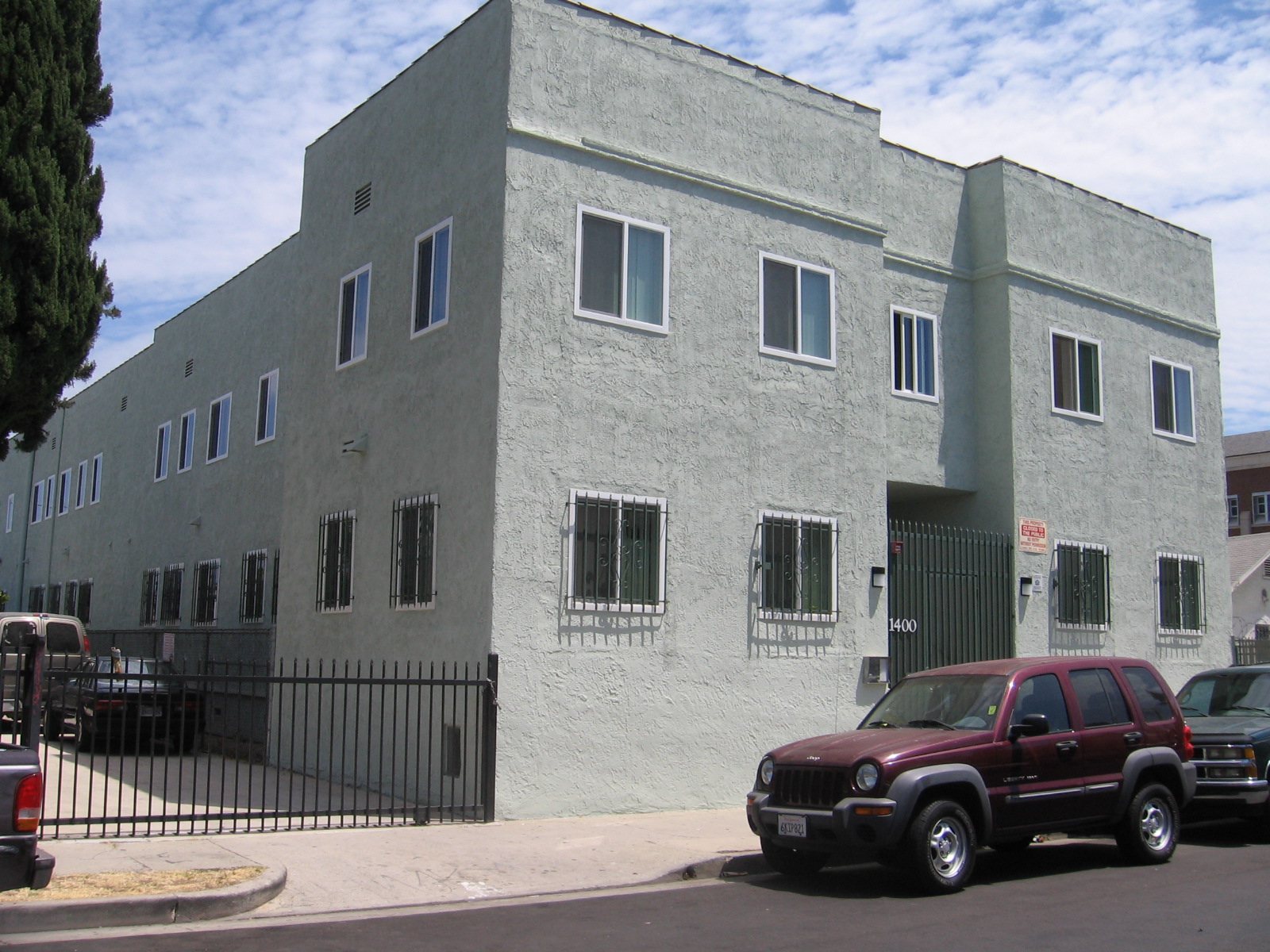 Left side view of a grayish green two story building, multiple windows with white trims, bottom windows with security bars, black iron locked-gate entrance, light lamps on each side, call box on the left side, building number on the left side of the gate