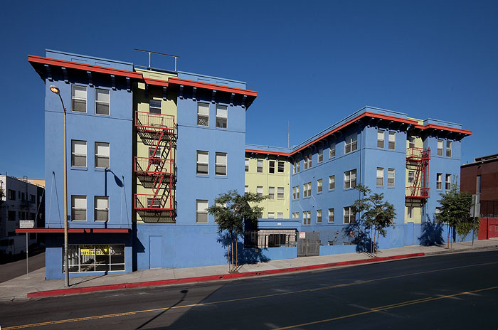 Front view of a four story blue building that sits on a slight hill. Entrance is gated. There are fire escapes in front of the building. There are three newly planted trees on the sidewalk.