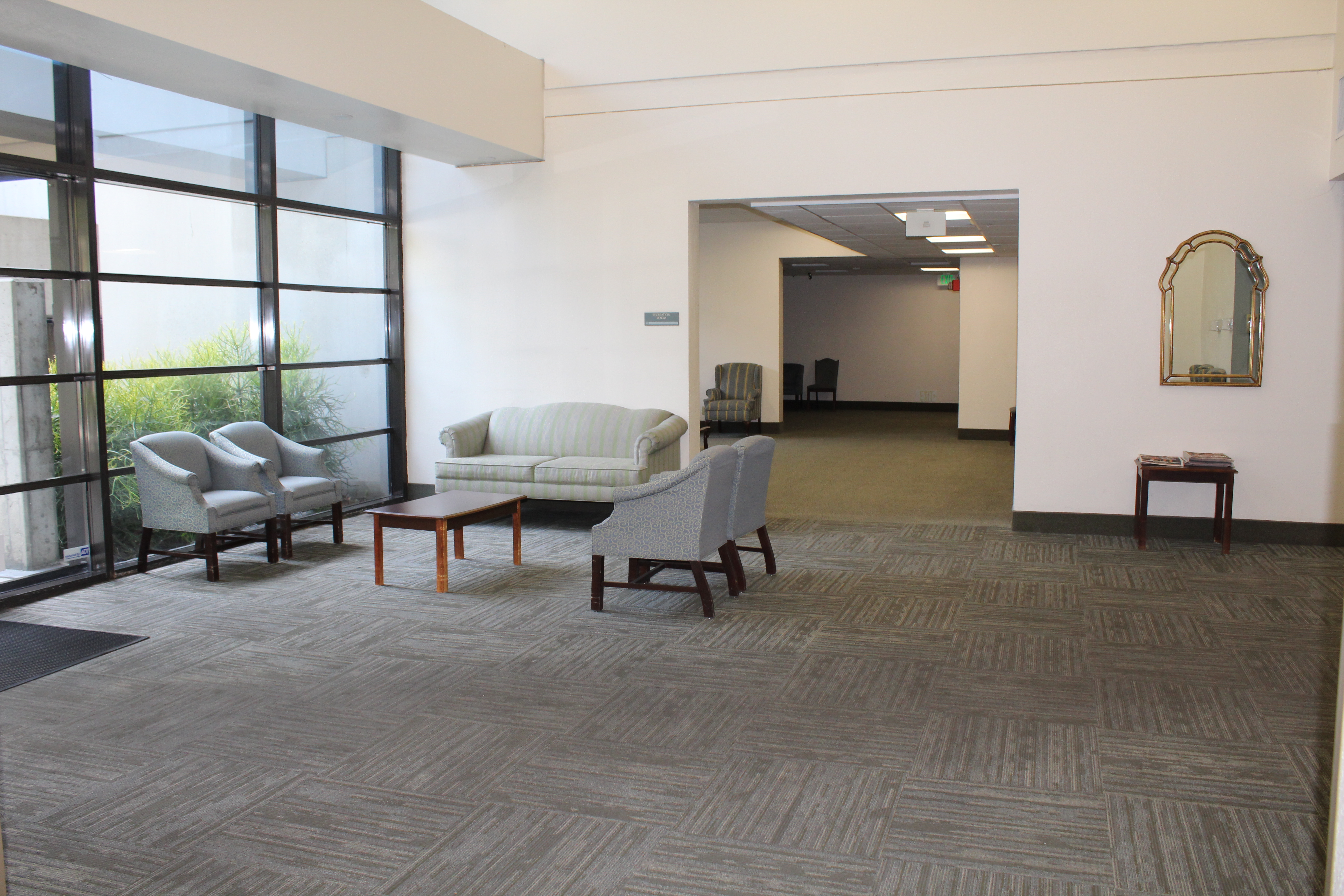 Interior view of a community room of Angelus Plaza 1. Grey sofa chairs, two across from each other with a rectangular coffee table inbetween them and a loveseat to the side next to entryway to another room.