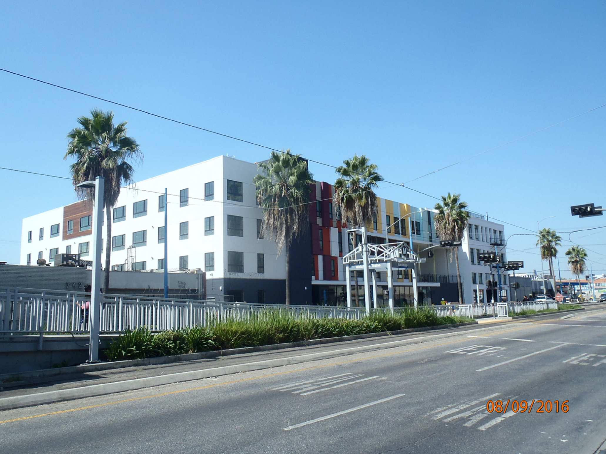 Four story white modern building with small stripes of red and yellow. Building is adjacent to a metrolink station. Entry way is ground level with tall windows. There are palm trees on the sidewalk in front of the building.