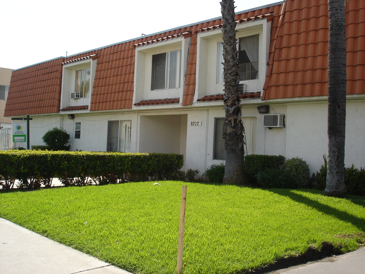 White 2 floor building with red roof tile, grass, bushes and trees in front area. Some units with a/c