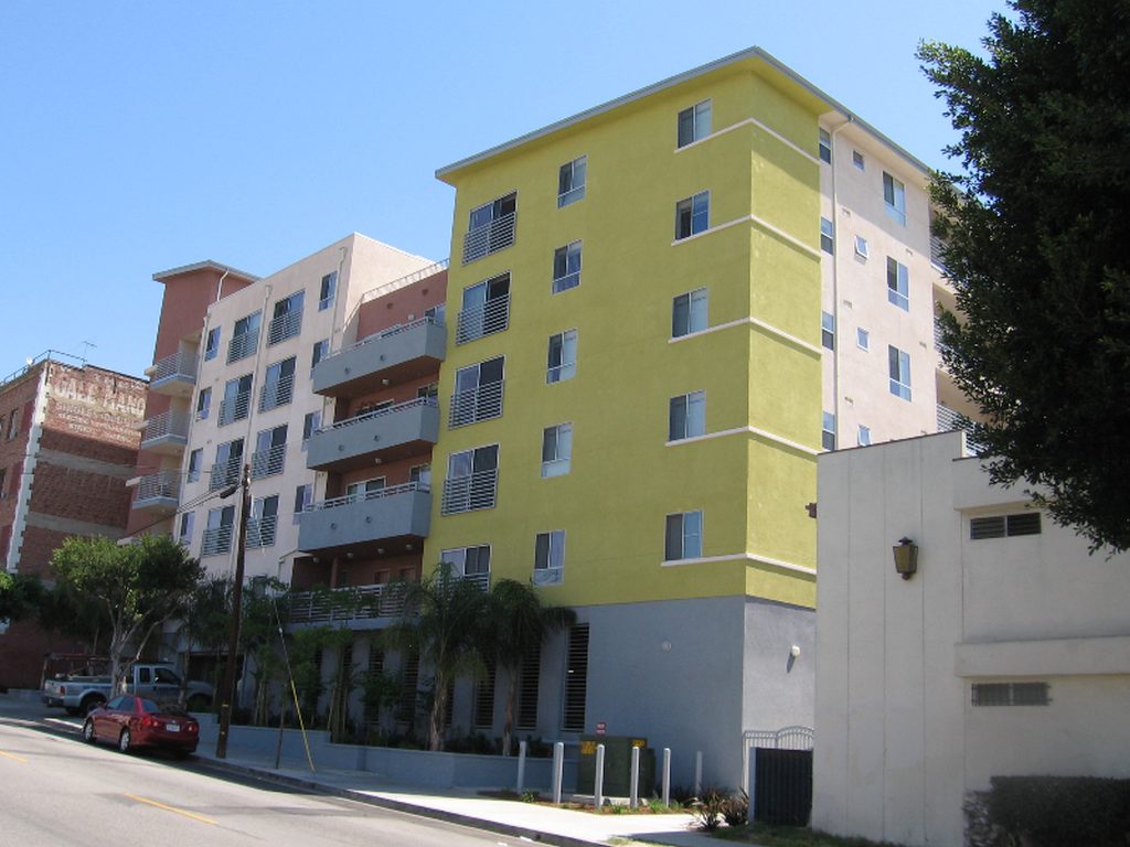 Five story multicolor building. Complex sits on a slight hill. Some units have balconies. Front of the building has a small area of varies trees and bushes.