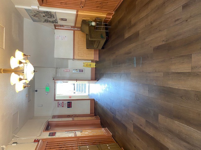 Different view of lobby area in which leads to an open door with a small staicase. There is a sofa seat on the corner with a small plant on the side, wood flooring, chandelier on the ceiling, and elevator and two metal doors on opposite sides.