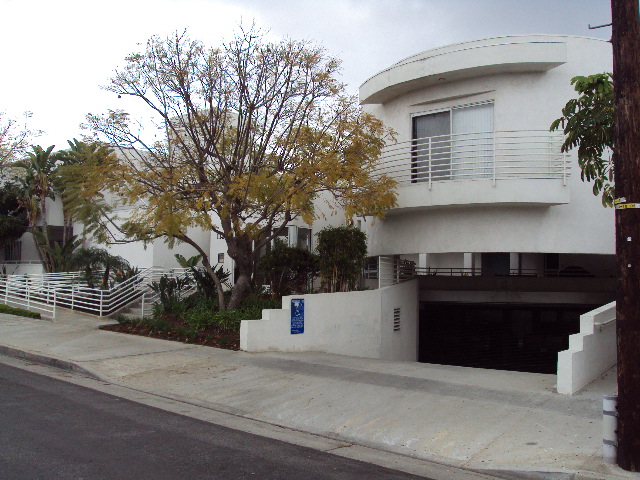 Right side of a white building, underground garage, top floor window with a white oval balcony, trees and plants all around the building.