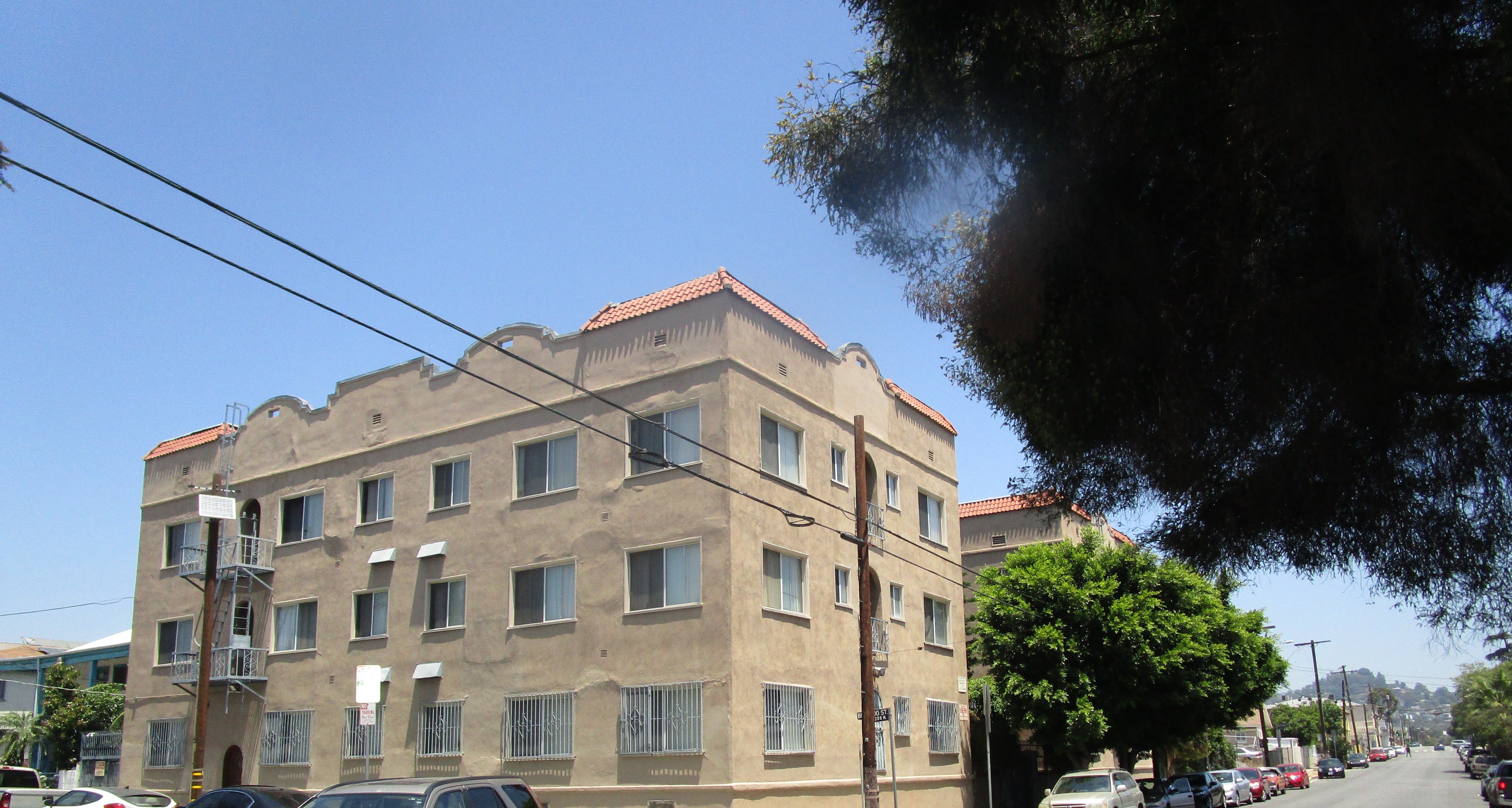 Corner view of the tan three floor building, multiple white windows, the bottom floor gated, Emergency stairs on one side, little brick style roof, to the right side part of the second building, two tall trees between the two buildings, light poles, cars