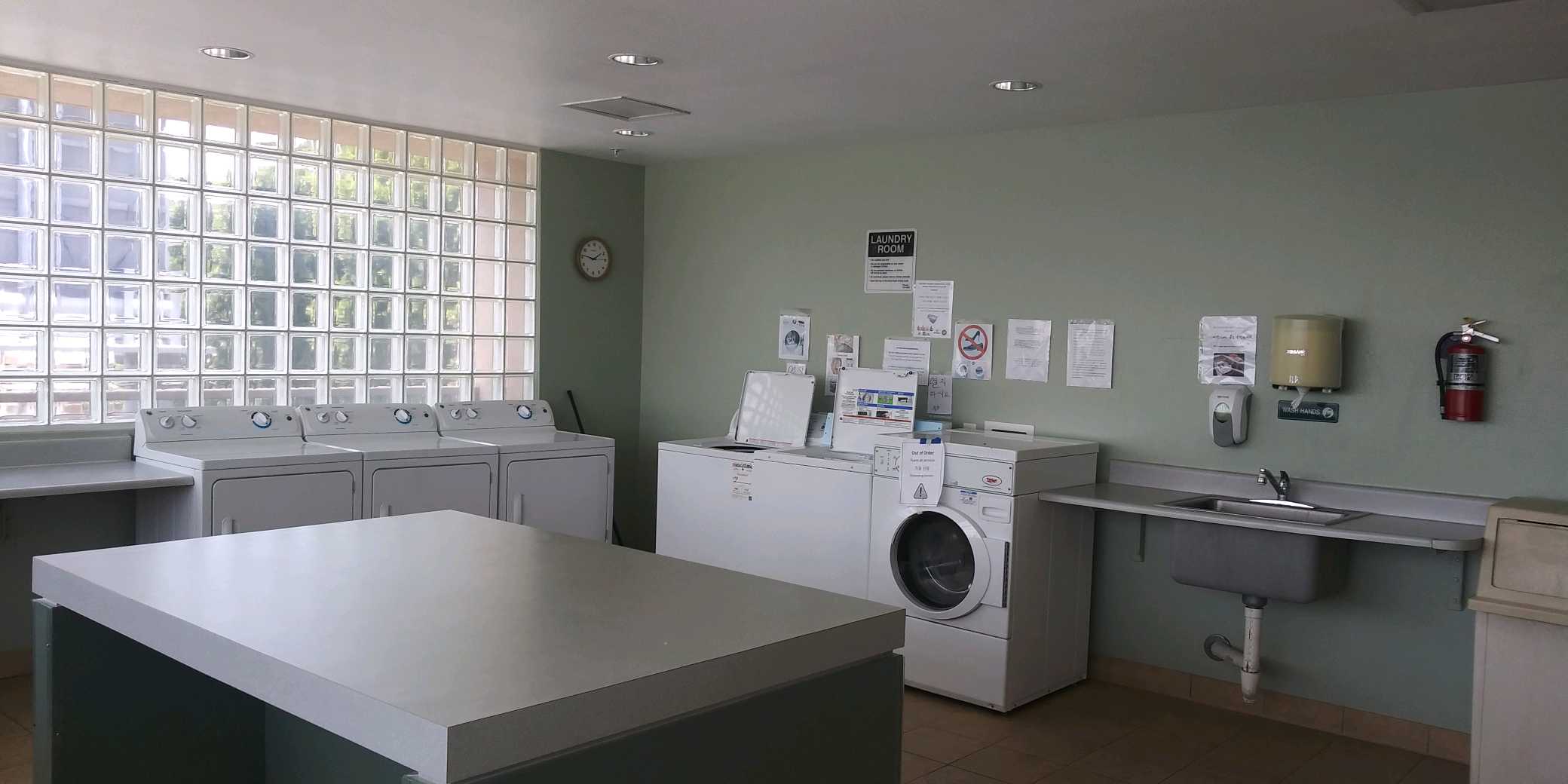 Laundry room with multiple washers and dryers. Center has a folding table. There is a large tiled window on one side of the wall. On the adjacent wall there are laundry room rules and signs posted. Thre is a sink, soap dispenser, paper towel dispenser, tr