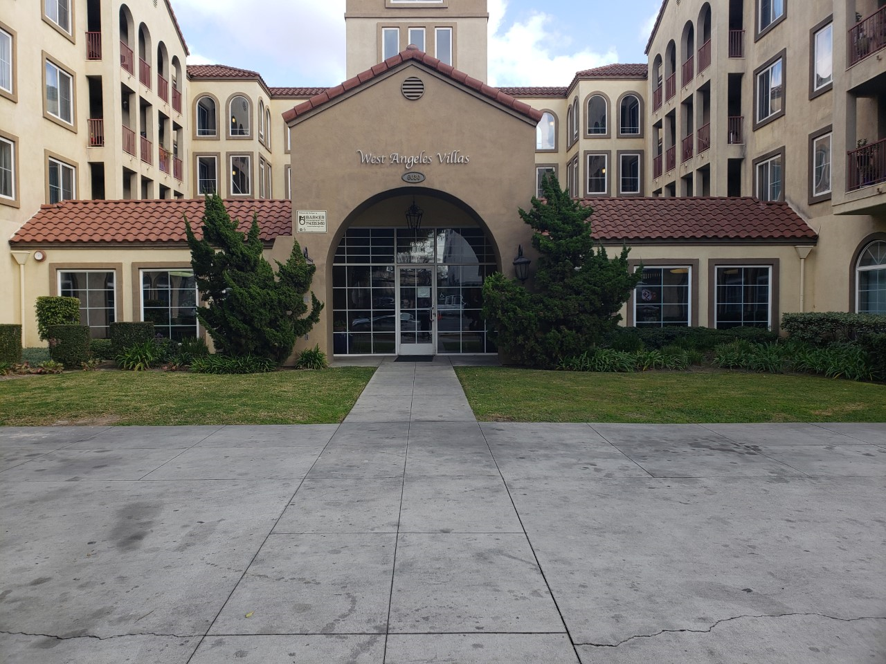 Front view of West Angeles villas. Wide walkway leading up to buildings front entrance. on either side of walkway there is a large area of grass with trees along building.