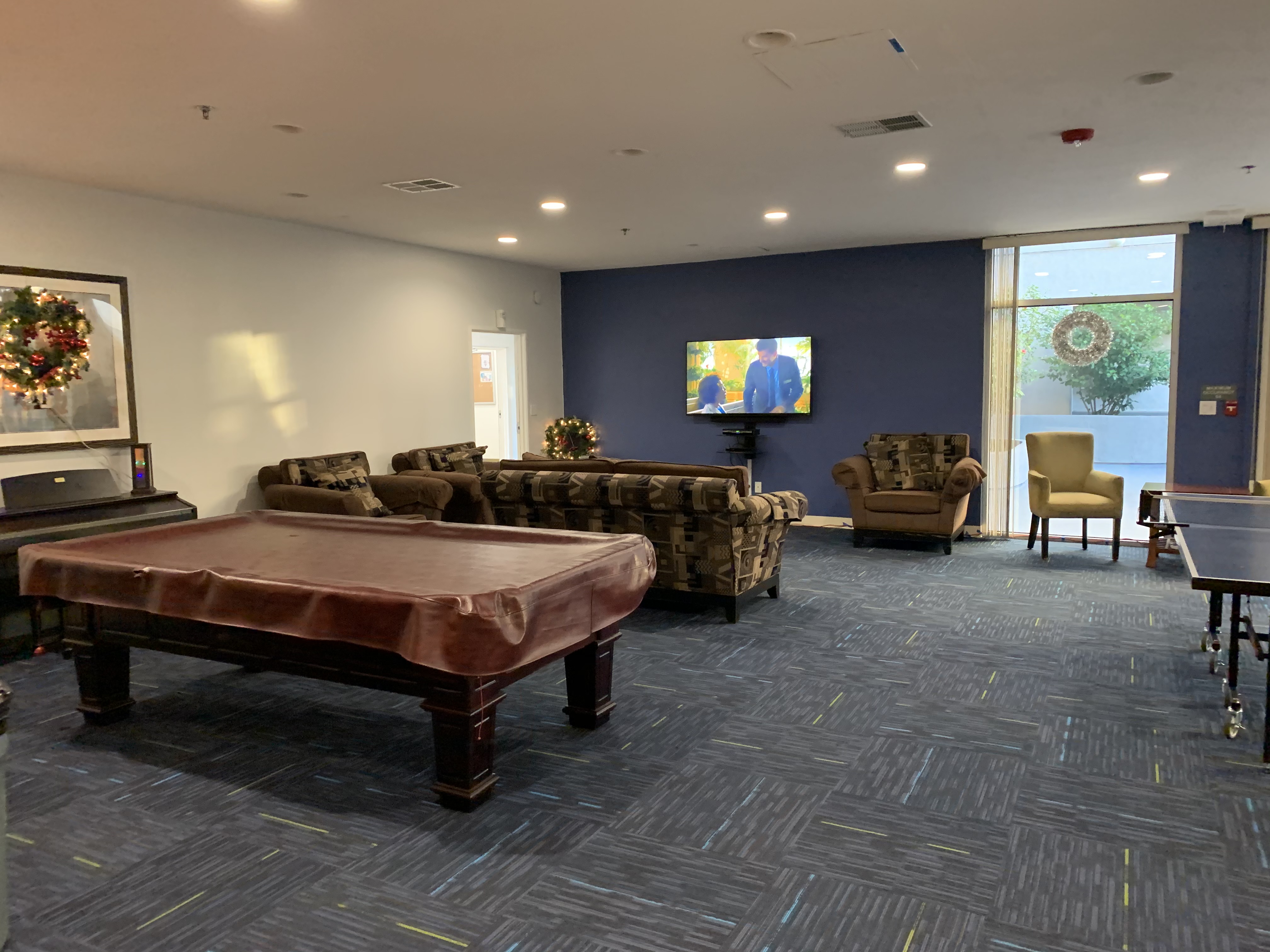 Community room that has a pool table, ping pong table, a piano, sofas, and a flat screen TV. There is a large window with vertical blinds.