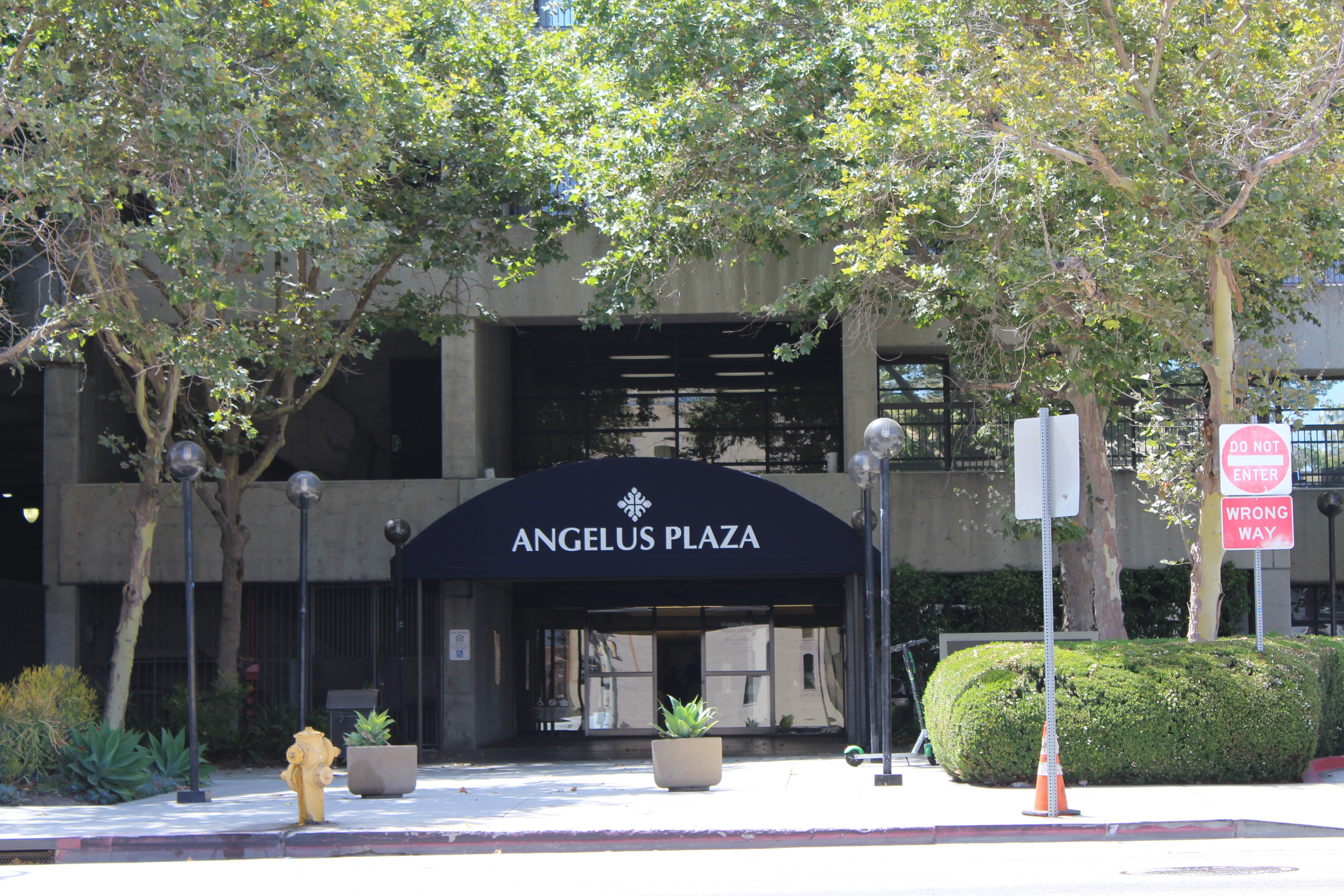 Street view of Angelus Plaza 1 entryway. Three lamp post on either side of the walk way with trees and bushes boarding it. Navy blue awning with white lettering