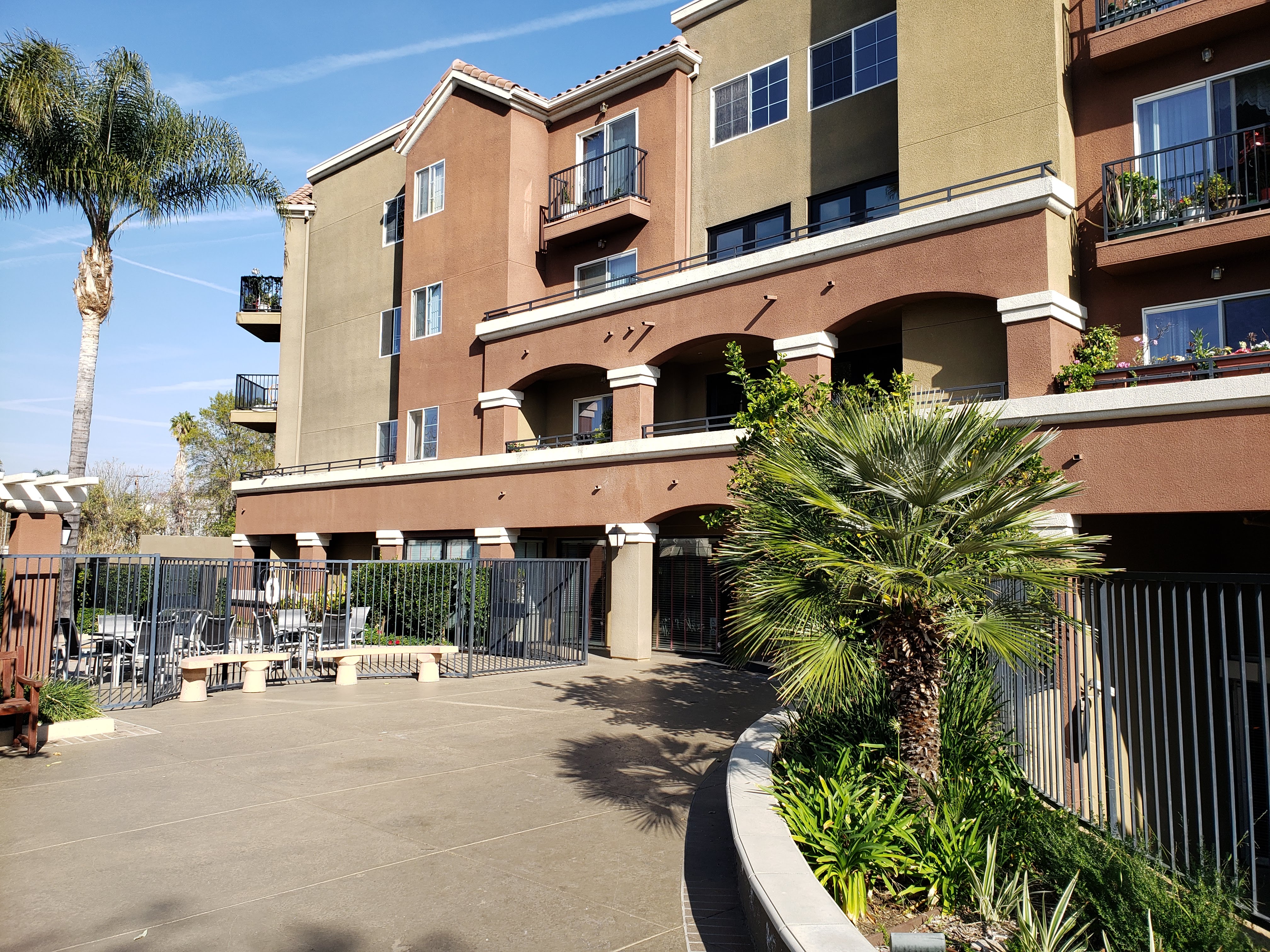 Image of vintage crossing senior apartments. Image includes gated outside spa area with pool side seating. large walkway with raised flower beds to right of walkway. walk way leads up to building. several balconys with flowers and miscellaneous objects pl
