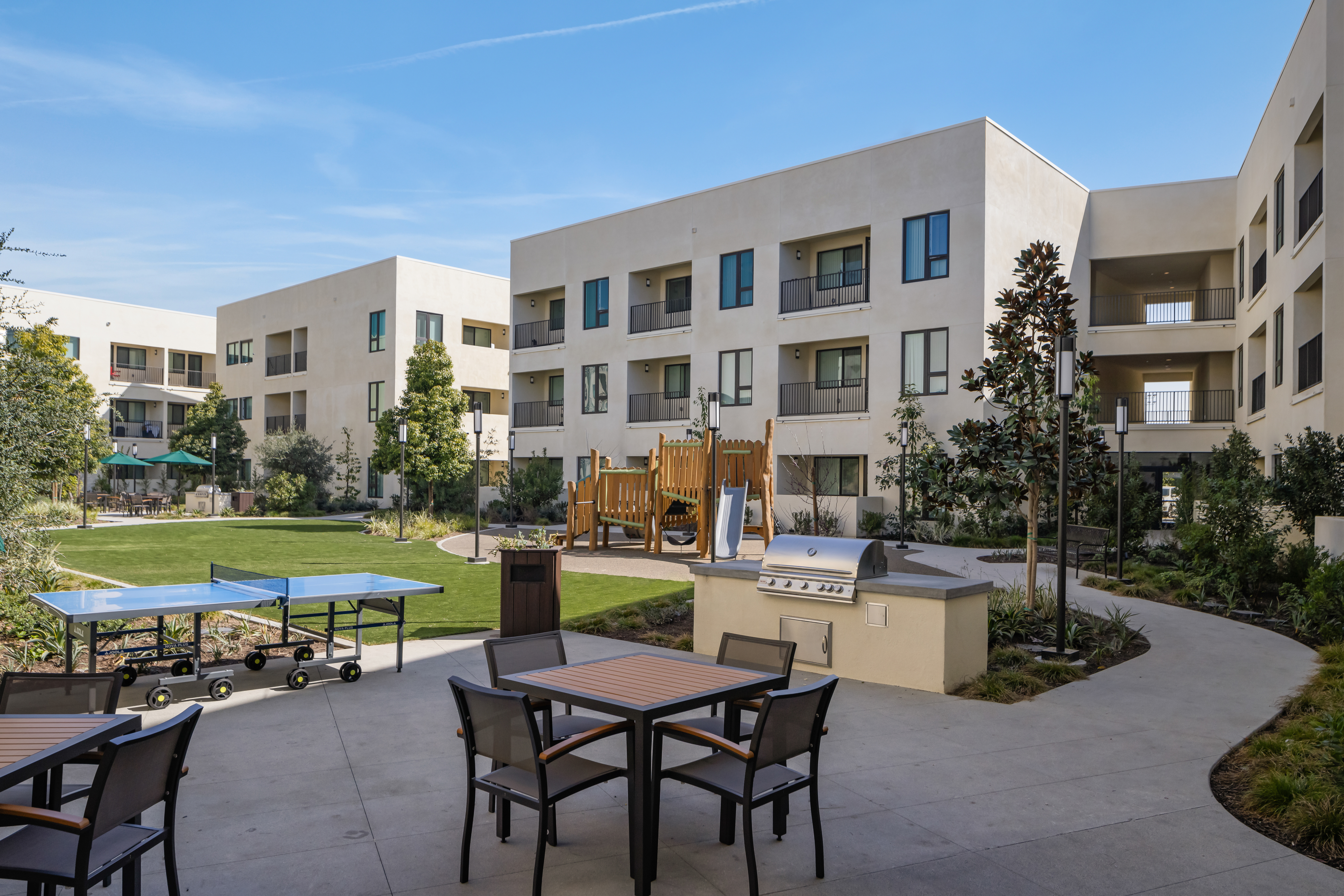 Modern apartment complex courtyard with tables, chairs, a ping pong table, a playground, a barbecue grill, and landscaped greenery under a clear blue sky.