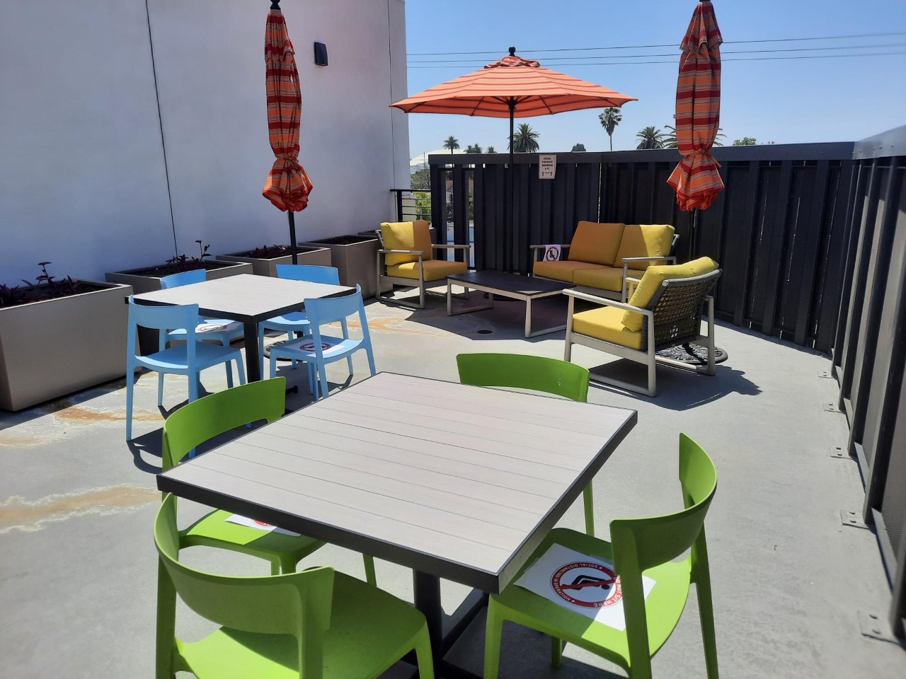 Outdoor deck area consisting of three tables, chairs, and sun umbrellas. This is surrounded by planters against a wall, and a fence closing off the area.
