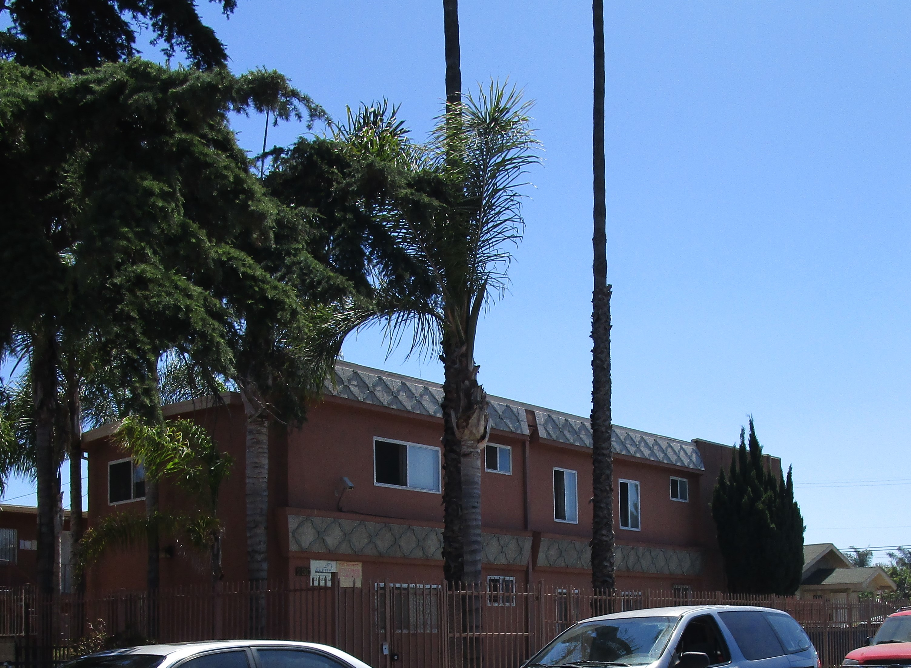 Side view of a brick color 2 story building, white and silver designs, gated, Property signs on the wall, a camera hanging, palm and big trees around the building, three cars parked in front.