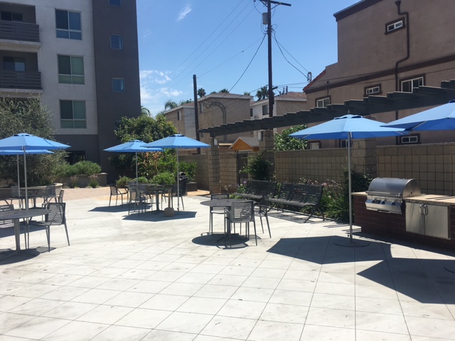 side view of a patio/courtyard, multiple metal tables with chairs and blue umbrellas, two metal benches, a Bar BQ grill, plants all over.
