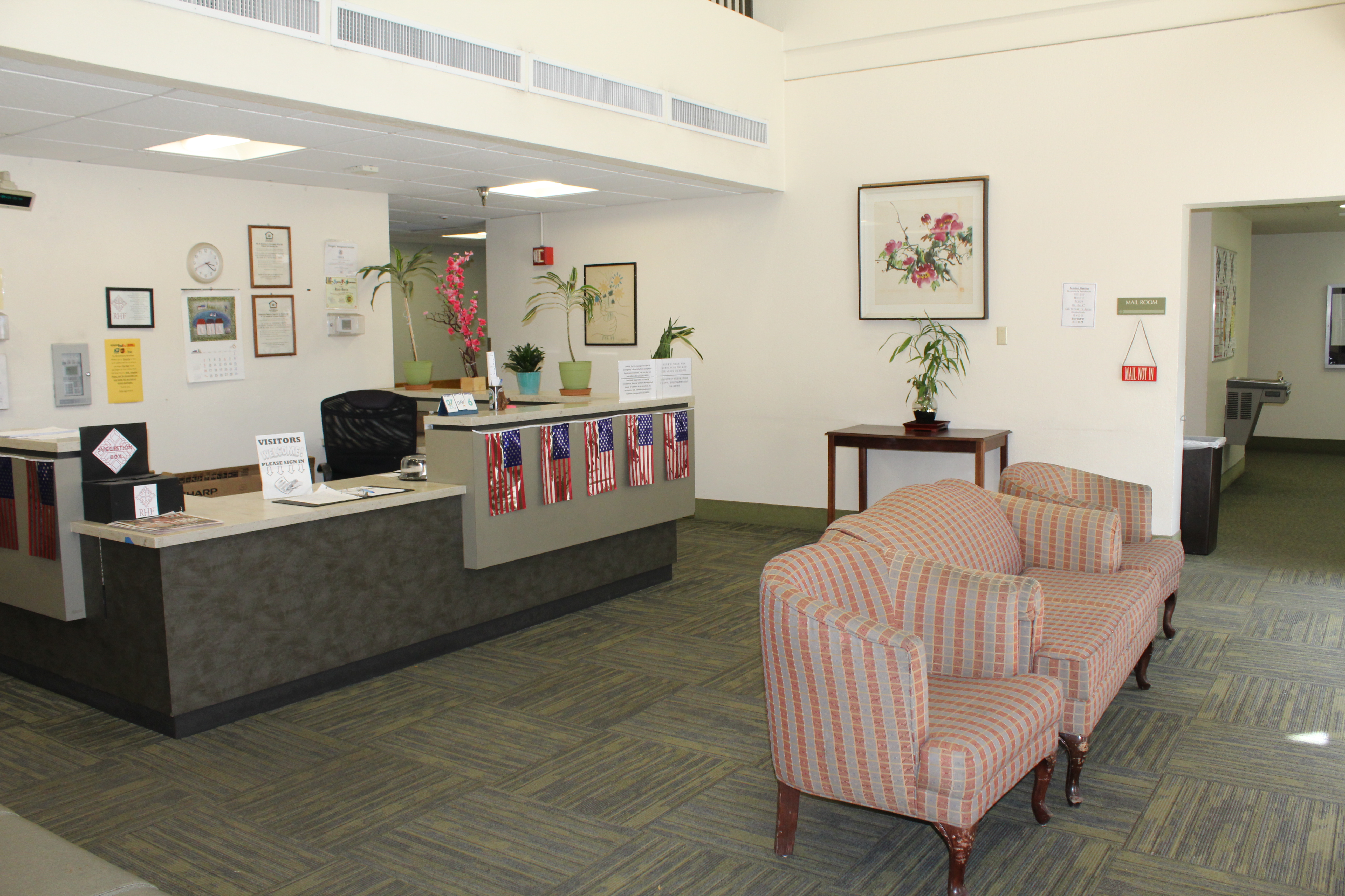 Interior view of a lobby area of Angelus Plaza 1. Potted plants around the front desk with American flag garland, 5 flags in all. Two sofa chairs with a love seat inbetween