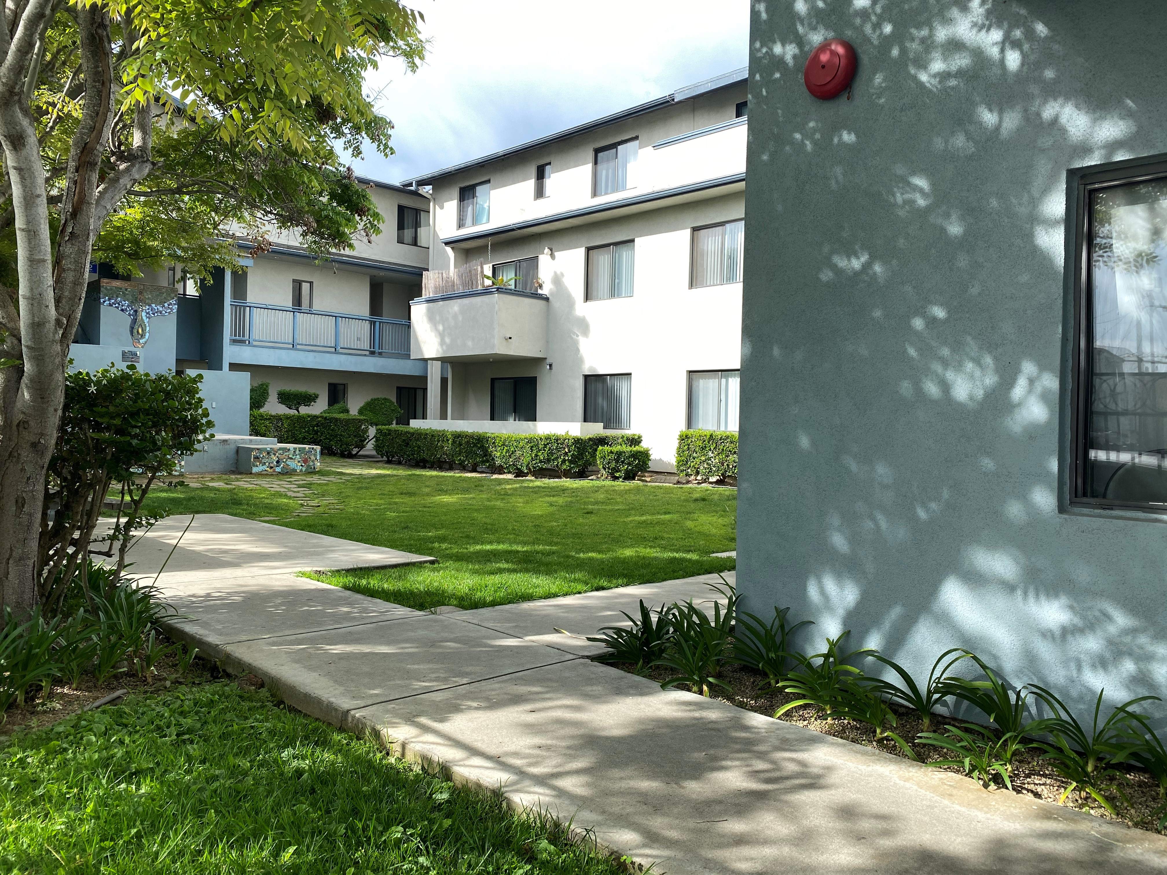 Courtyard of a three story building. There is a open grass area with adjacent bushes and trees. There is a cement passageway that leads to the units.