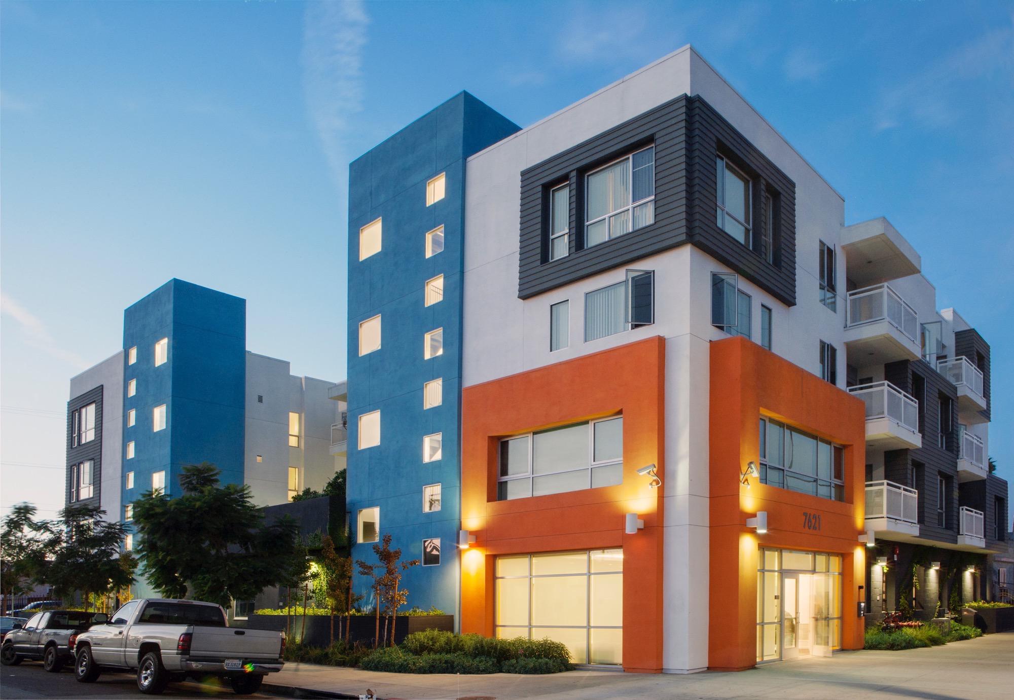Front side view of a modern four story building. Buildong colors consist of light blue, white, gray and orange. Some units have tall windows, and some have balconies. There is newly planted trees in the front sidewalk and bushes on the side of the buildin