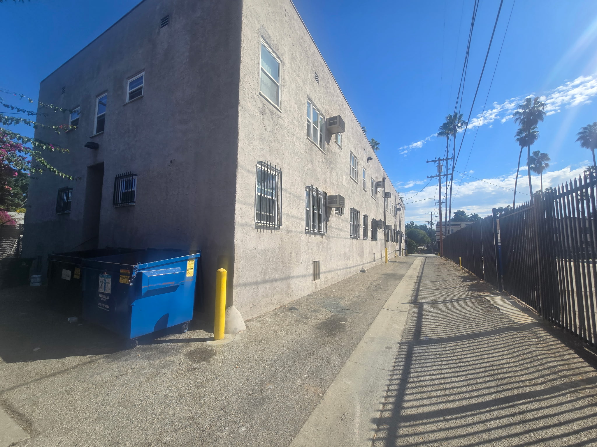 View of rear exterior of building, showing adjacent alley way,  building with metal bars on windows and large trash bins at exterior. 