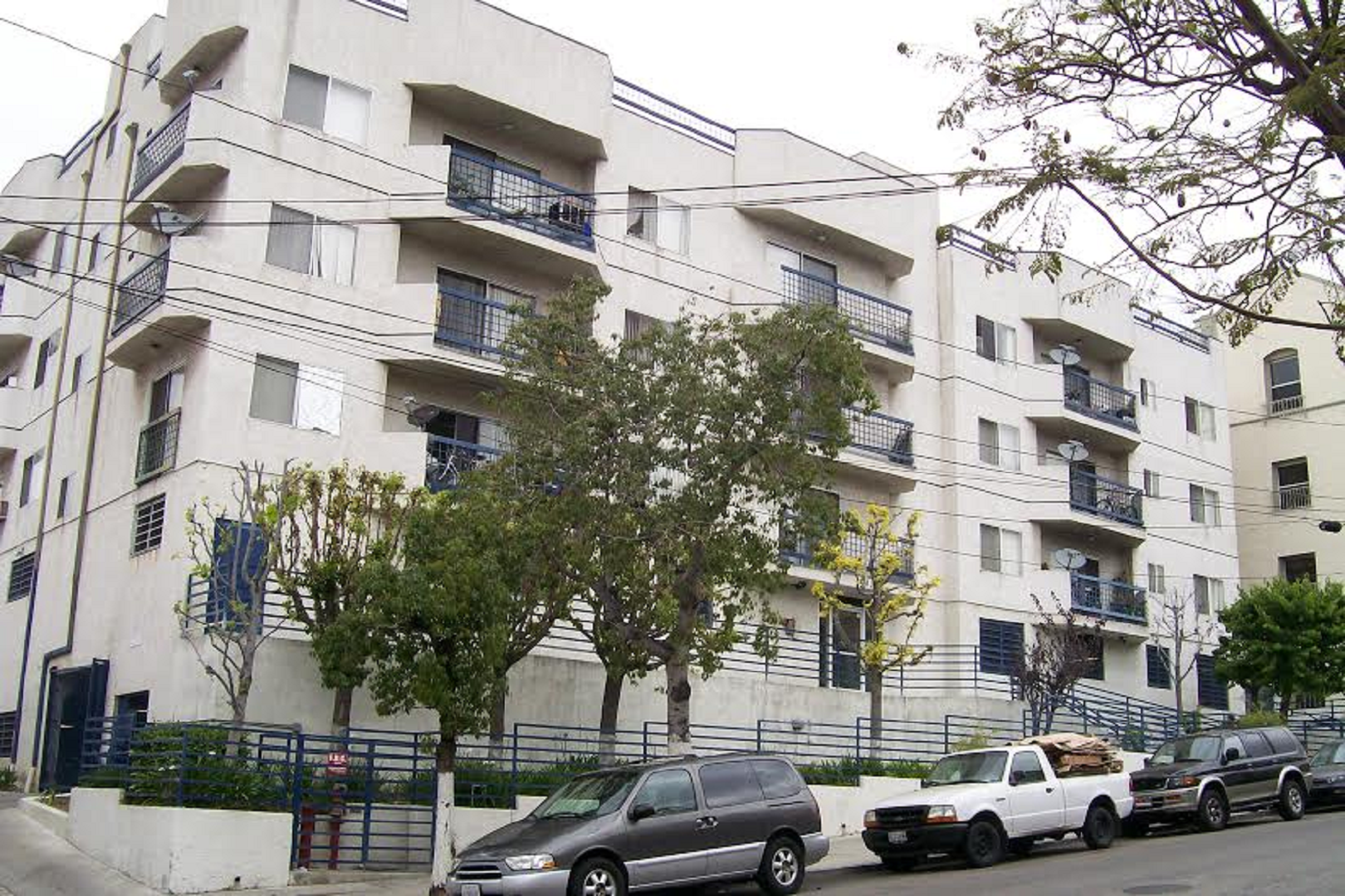 Five story white building that sits on a hill. Front entrance has blue long bars.Building also contains balconies with blue bars.