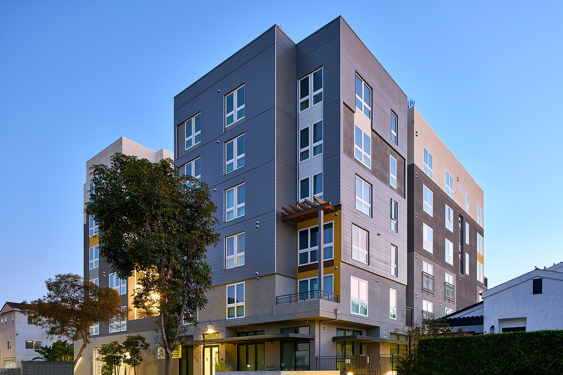A six story, mid-rise apartment building with gray panel siding, large windows, and balconies, photographed at dusk with a tree in front.