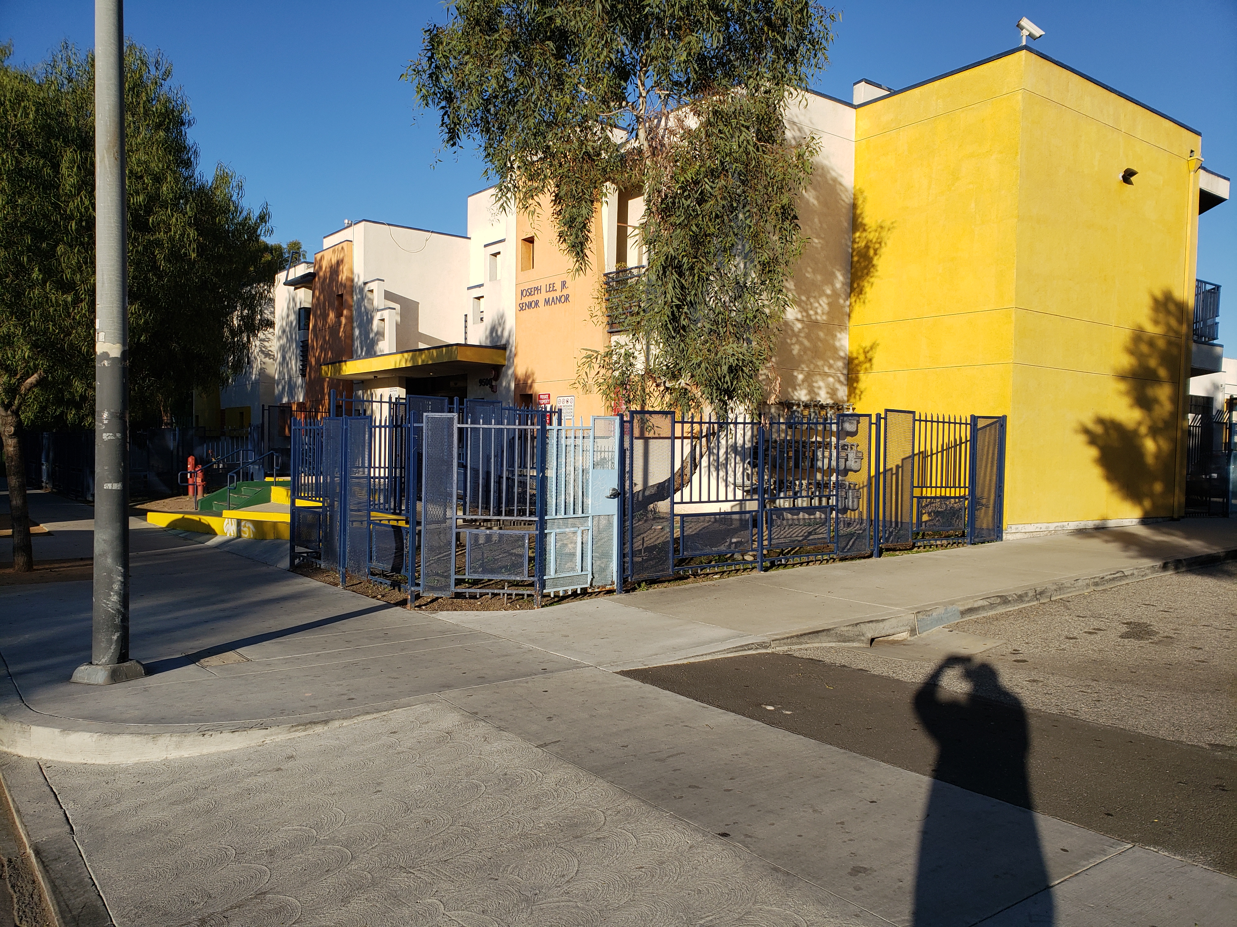 Street view of Heavenly Vision Seniors Apartments, Yellow and White building with with a blue gate and a tall tree inside the gate