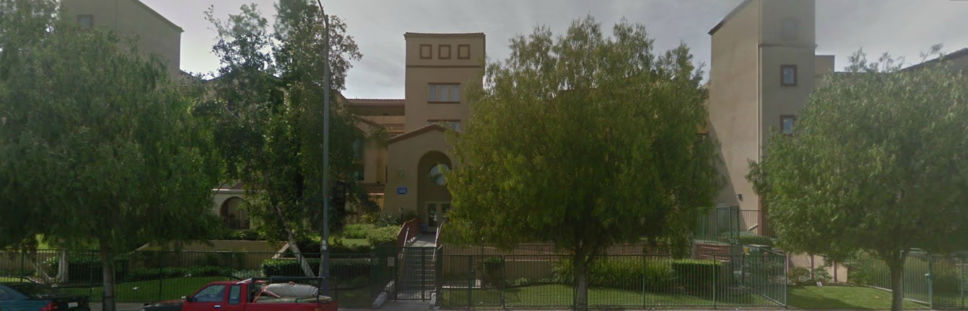 Front view of large light brown building. Front is gated. There is a grass area behind the fence with with bushes and a tree. There are stairs that lead to the building's entrance with handlebars on both sides. Three trees sit on the sidewalk in front of