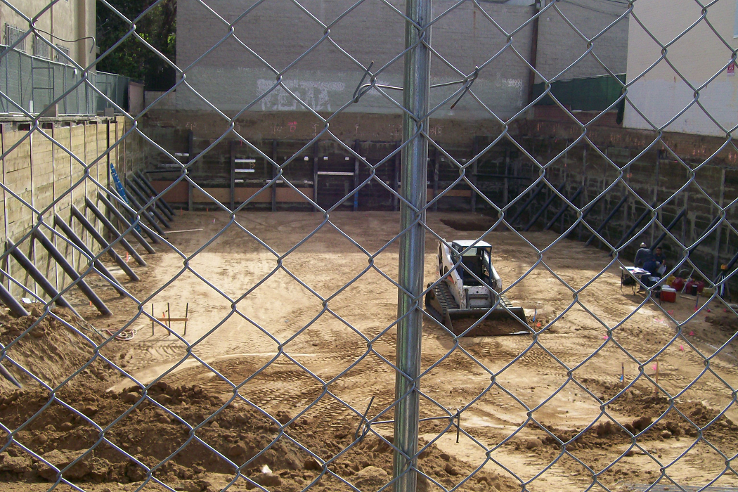 Zoom in of a fenced construction site, a white bulldozer, construction materials, sticks with colorful flags, two males talking, dirt.