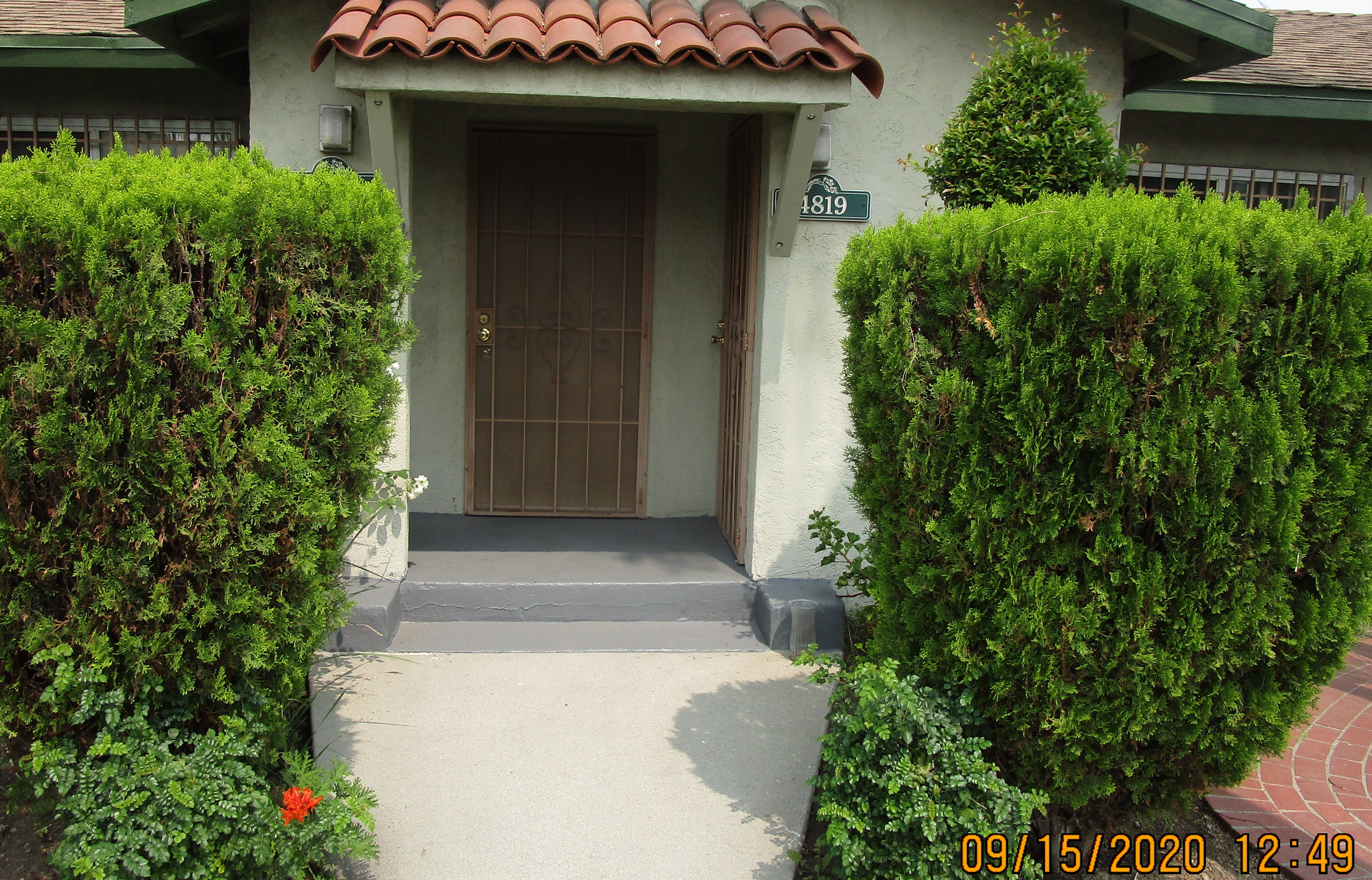 Front view of main entrance, one step, another door on the right side, house number on the right side, big tall bushes on each side.