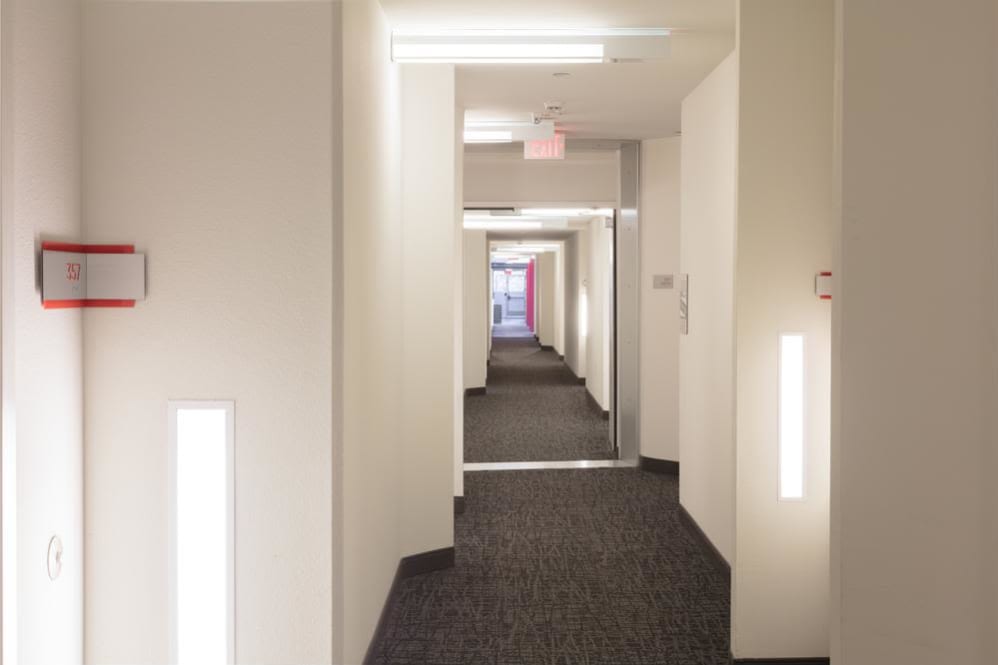 Inside view of a hallway that leads to multiple units. It is well lit and there is an exit sign. Floor is carpeted.